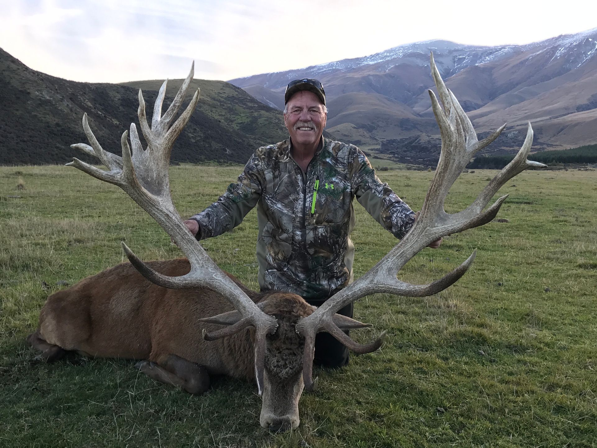 A smiling man in camouflage kneels next to a large red deer stag with impressive antlers on a grassy field in front of mountains.