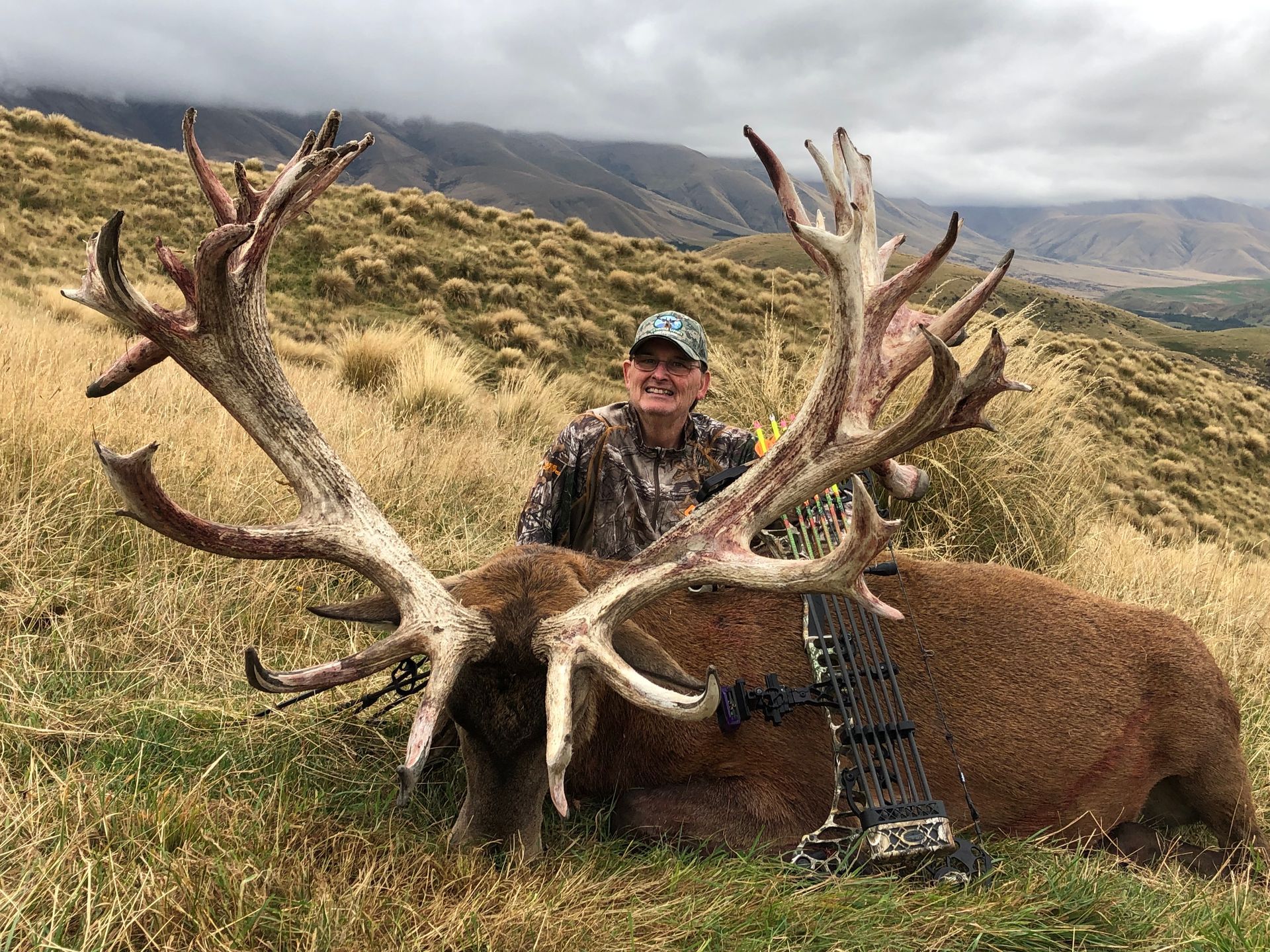 Man in camouflage with a large dead red deer. The deer has massive antlers; they and the man are in a grassy hillside setting.