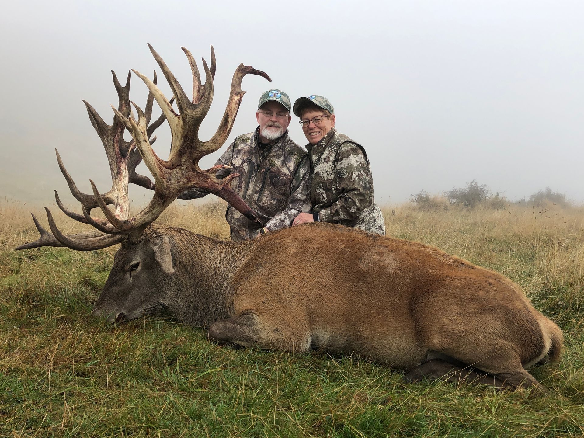 Two people in camouflage stand over a large, deceased red deer with impressive antlers in a grassy field; a foggy background.