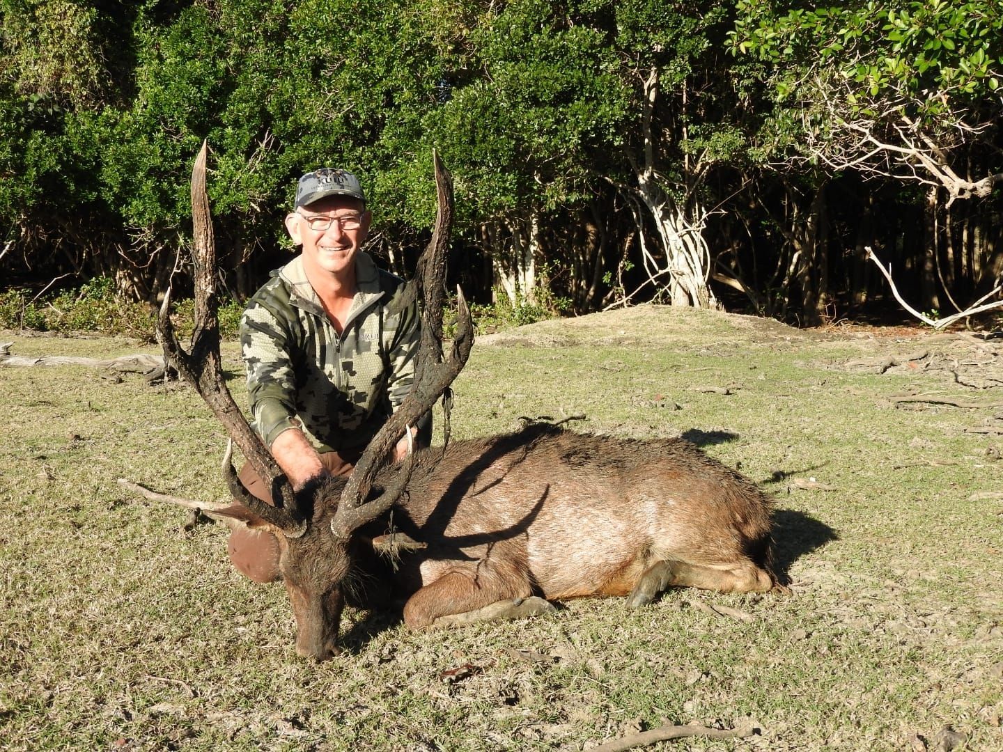 Man in camouflage smiles next to a dead stag with large antlers on grass; trees in the background.