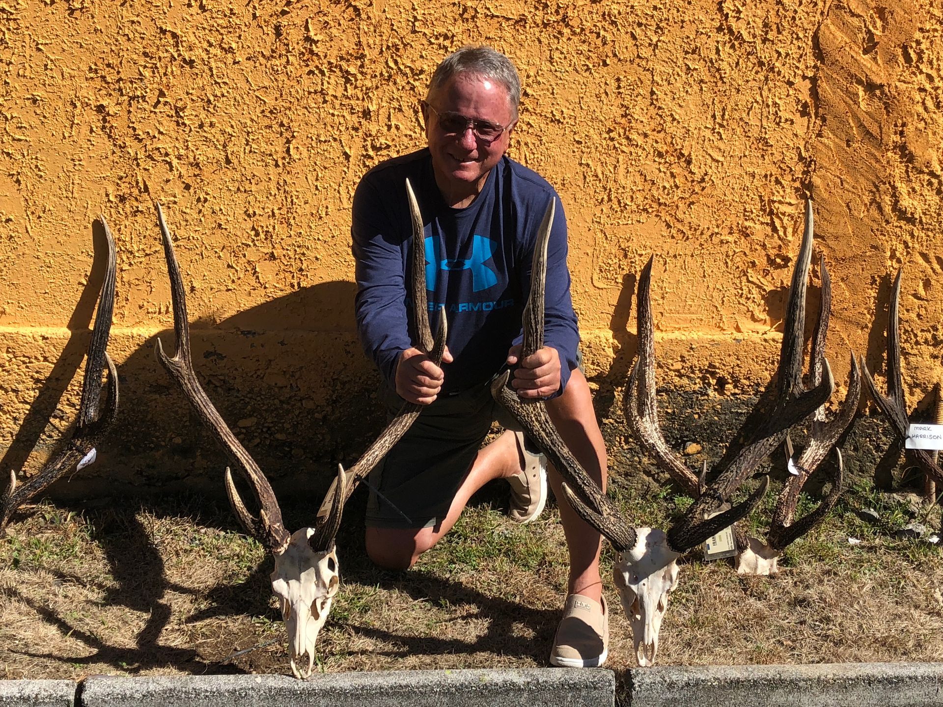 Man kneeling outdoors, holding antlers, surrounded by deer skulls and antlers against an orange wall.