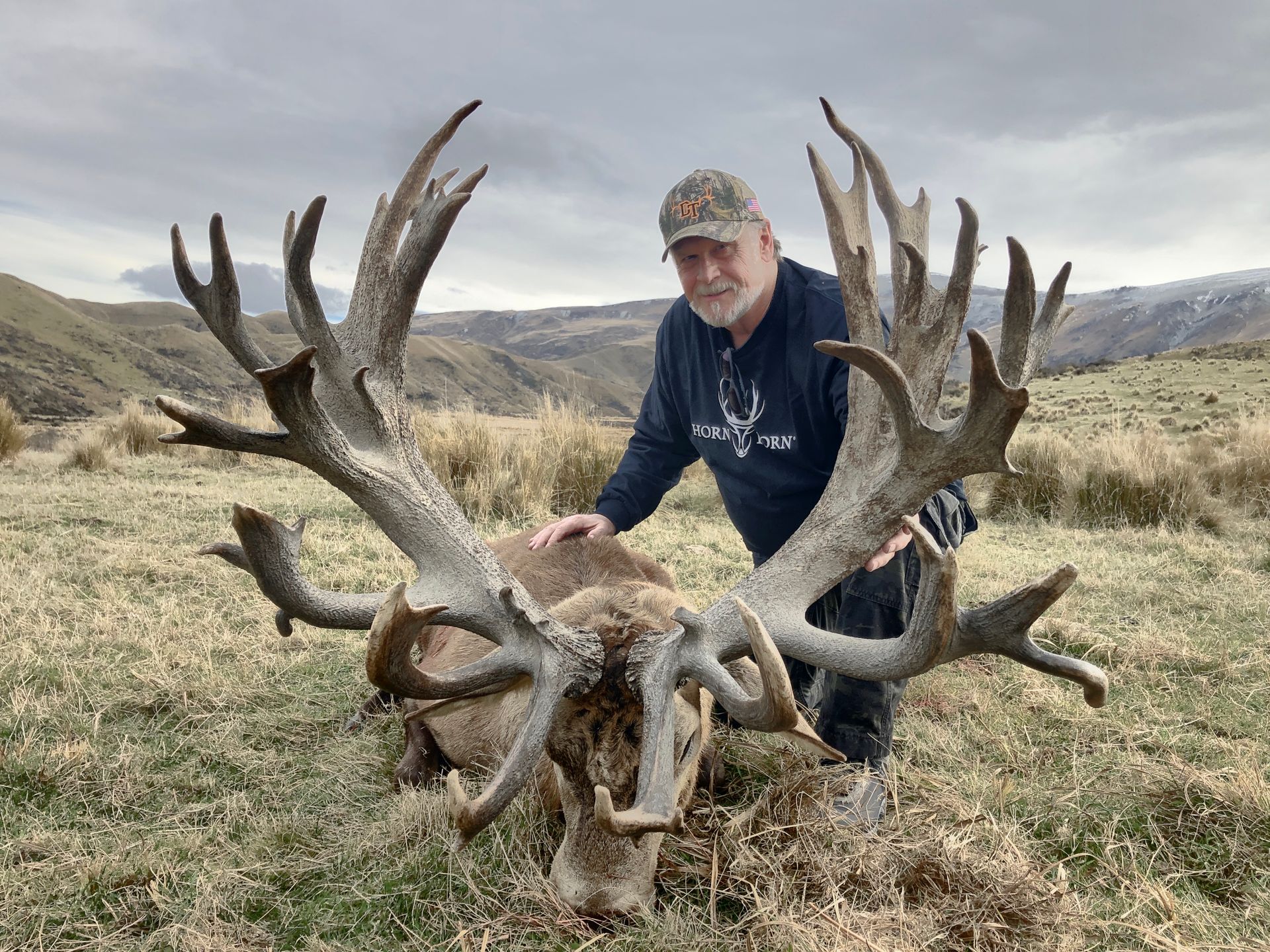 Man in camo hat poses beside a large dead deer with massive antlers on a grassy hillside.