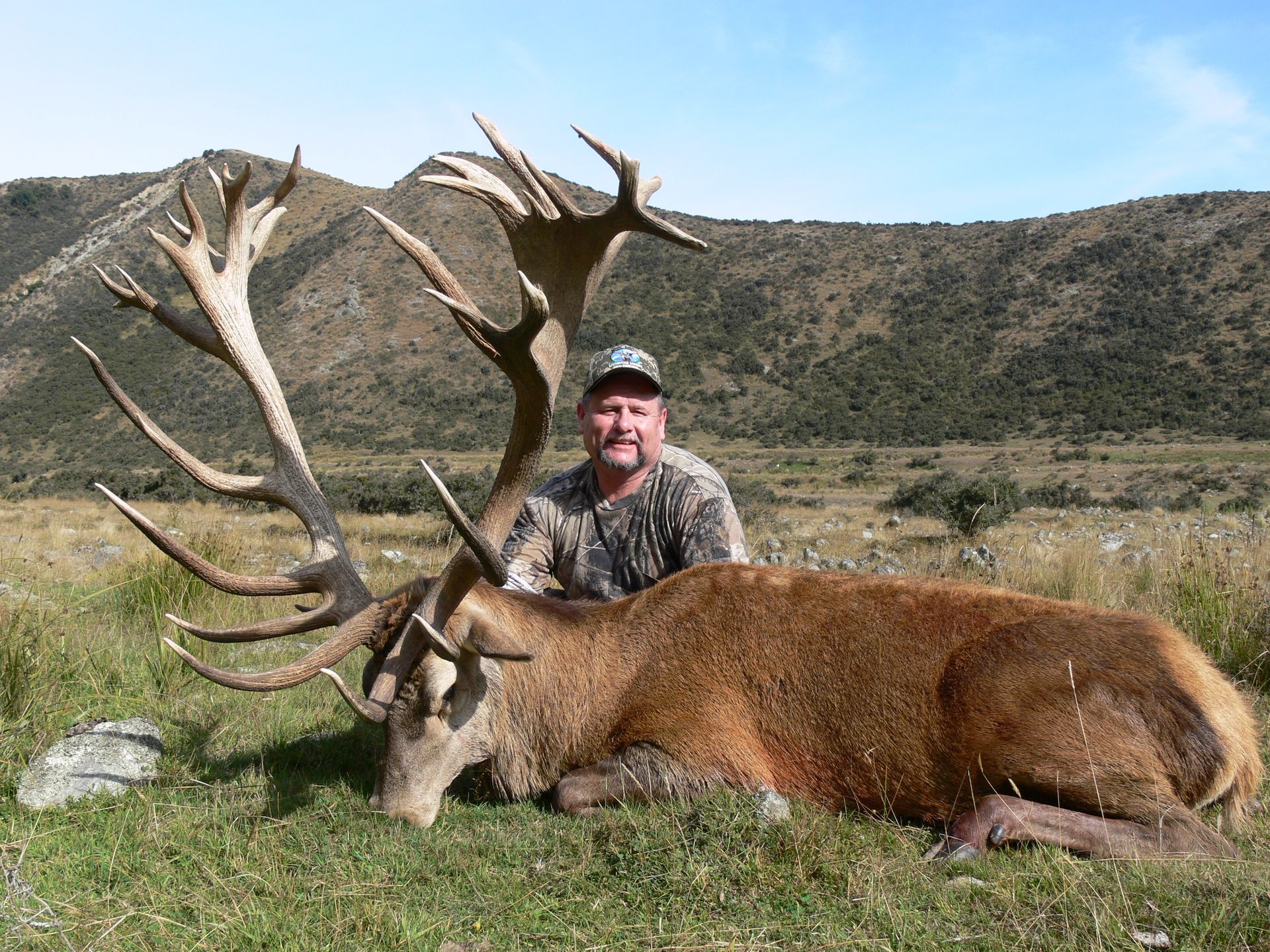Man in camo posing with a large, red deer carcass with impressive antlers in a grassy, mountainous landscape.