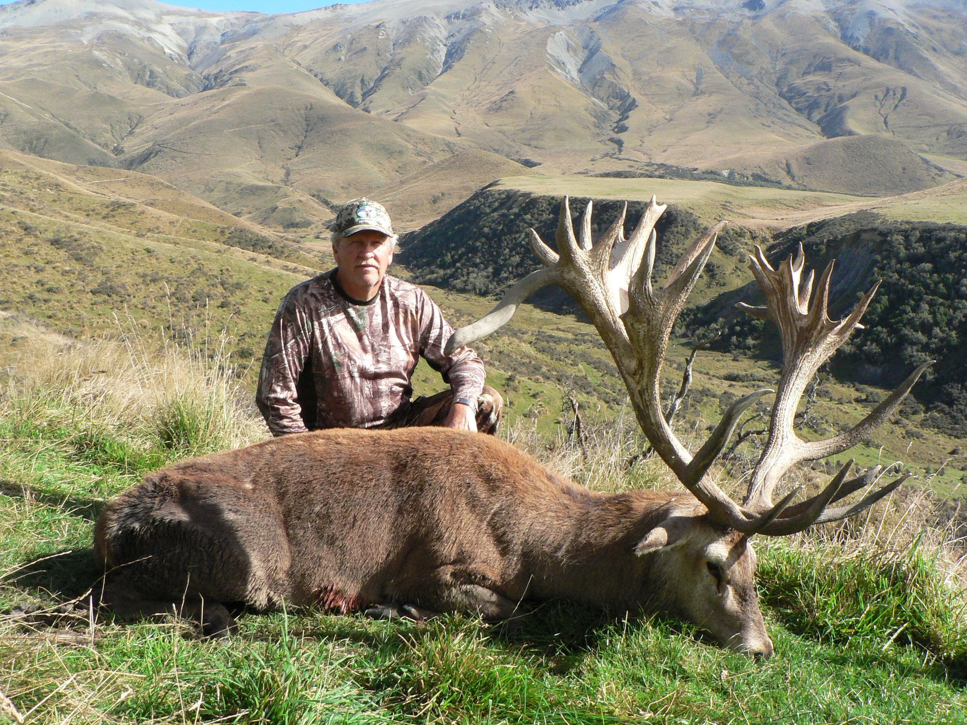 A man in camouflage kneels beside a large red deer with impressive antlers in a grassy mountain landscape.