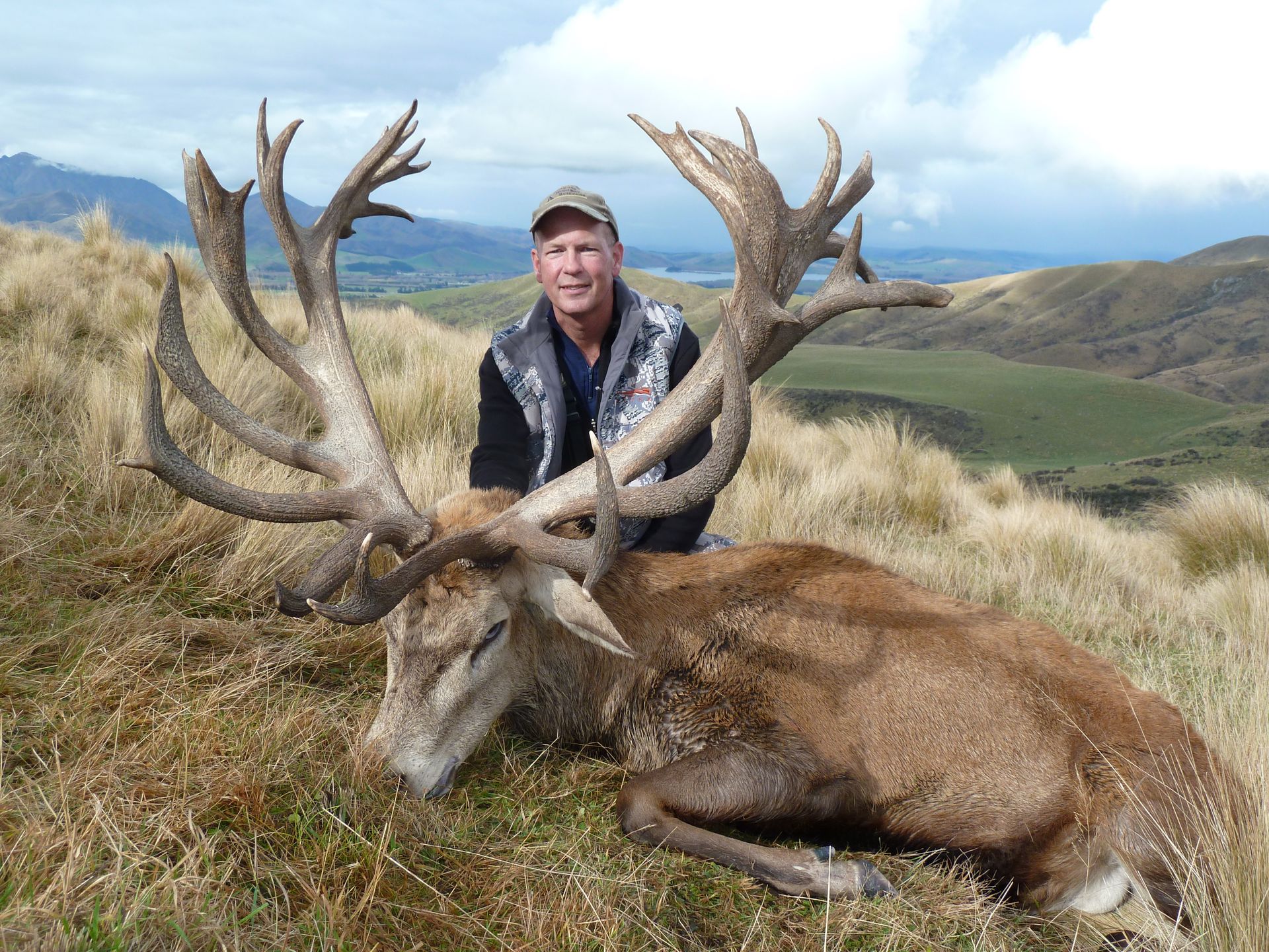 Man poses proudly with a large, deceased red deer with impressive antlers in a grassy, mountainous setting.