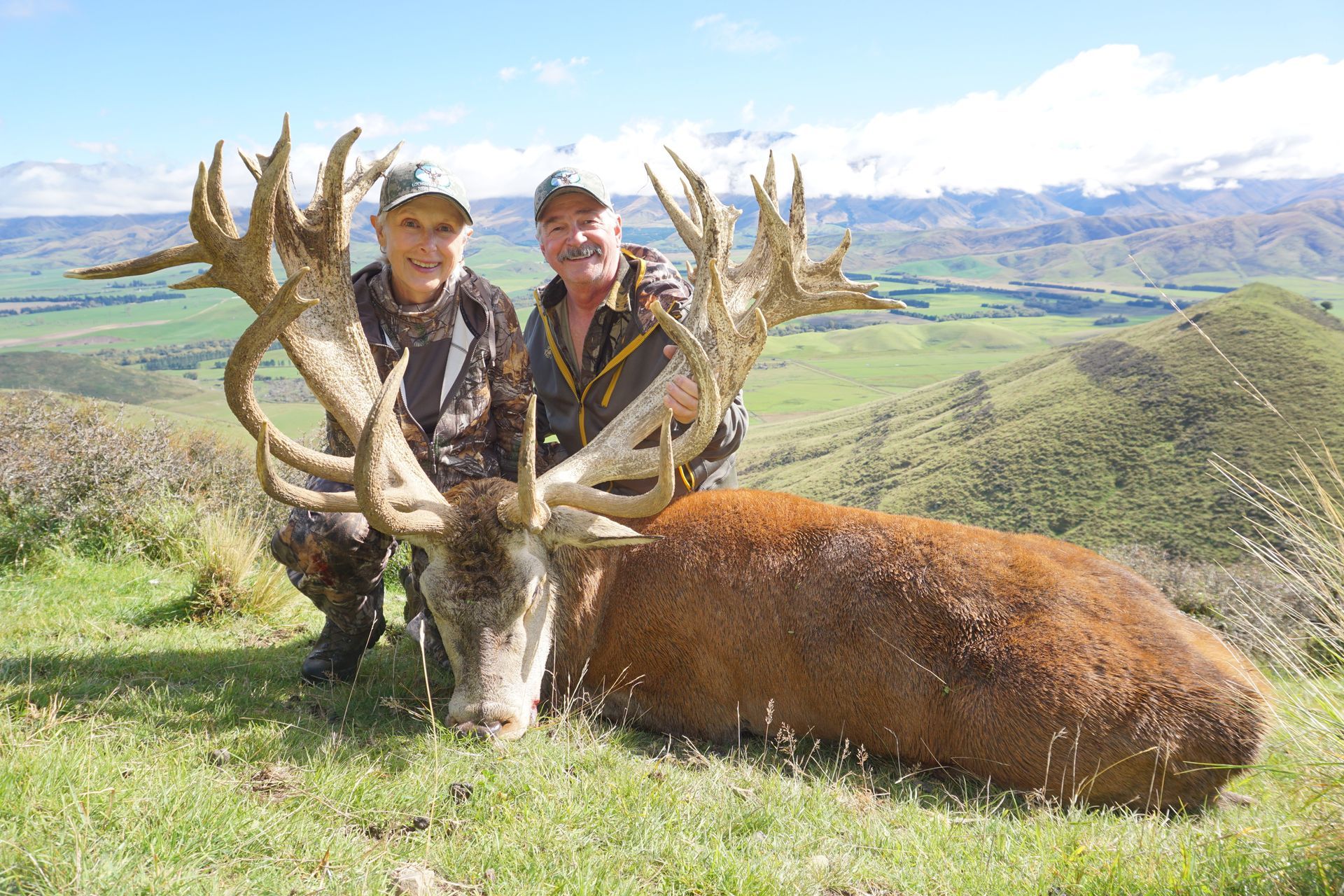 Two people kneeling beside a large dead red deer with impressive antlers, green hills, and snowy mountains in the background.