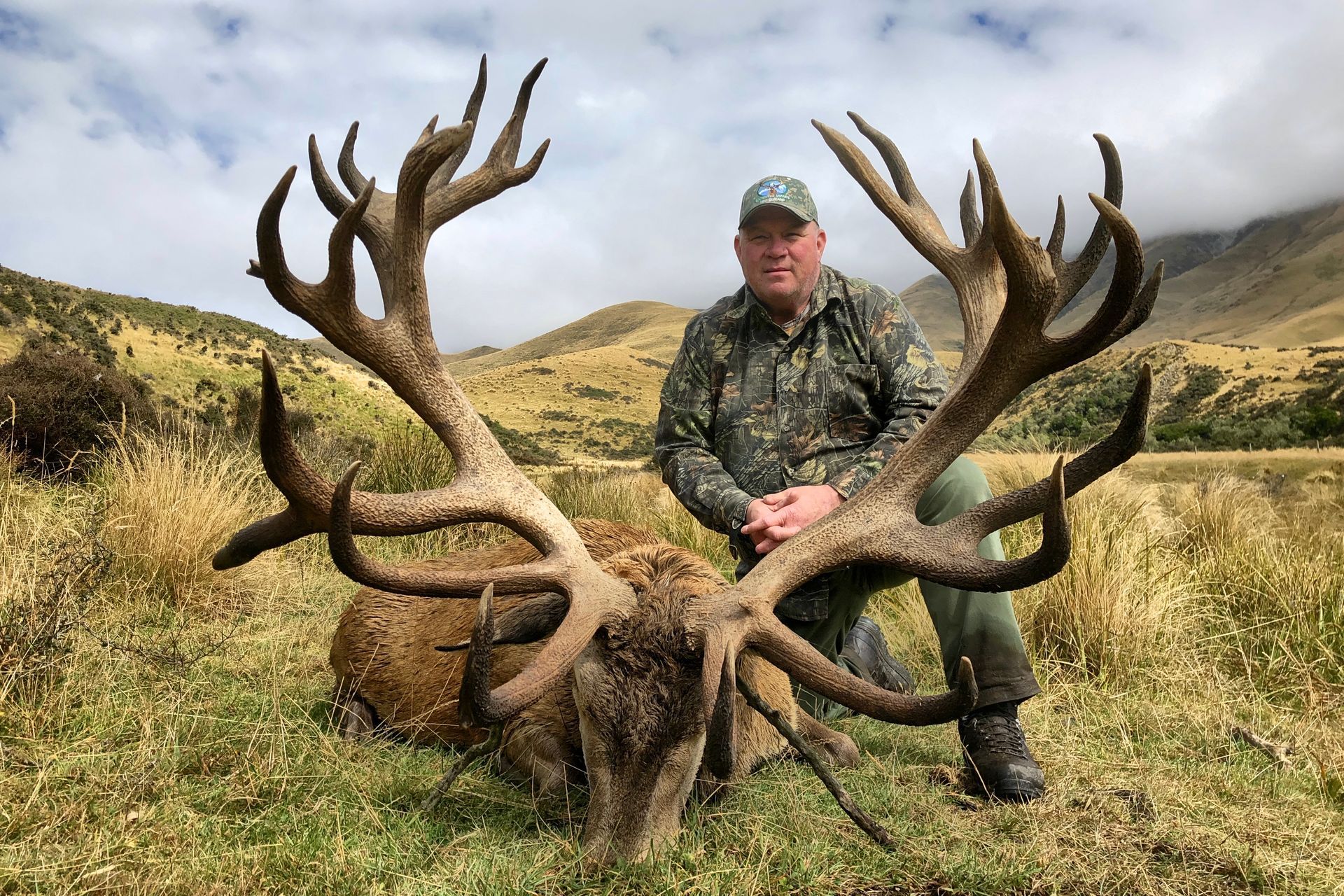 Man kneels beside a large dead deer with huge antlers in a grassy, mountainous landscape. Man wears camouflage and a hat.