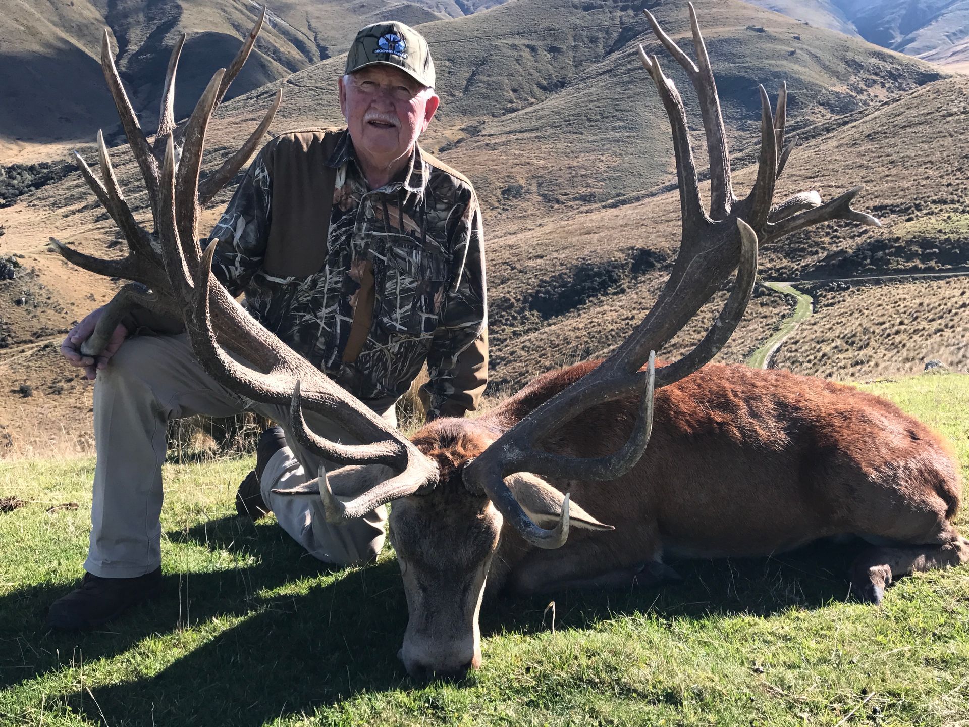 Man kneels beside a large, red deer with impressive antlers, outdoors in a grassy, mountainous setting. The man smiles, dressed in hunting attire.