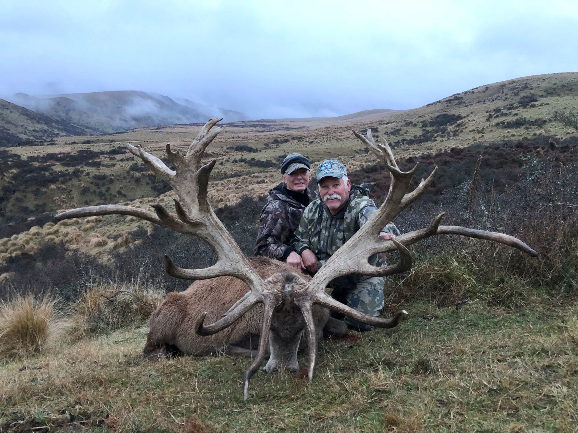 Two people pose with a large dead deer, its antlers spread wide, against a hilly, overcast backdrop. Both are wearing camouflage.