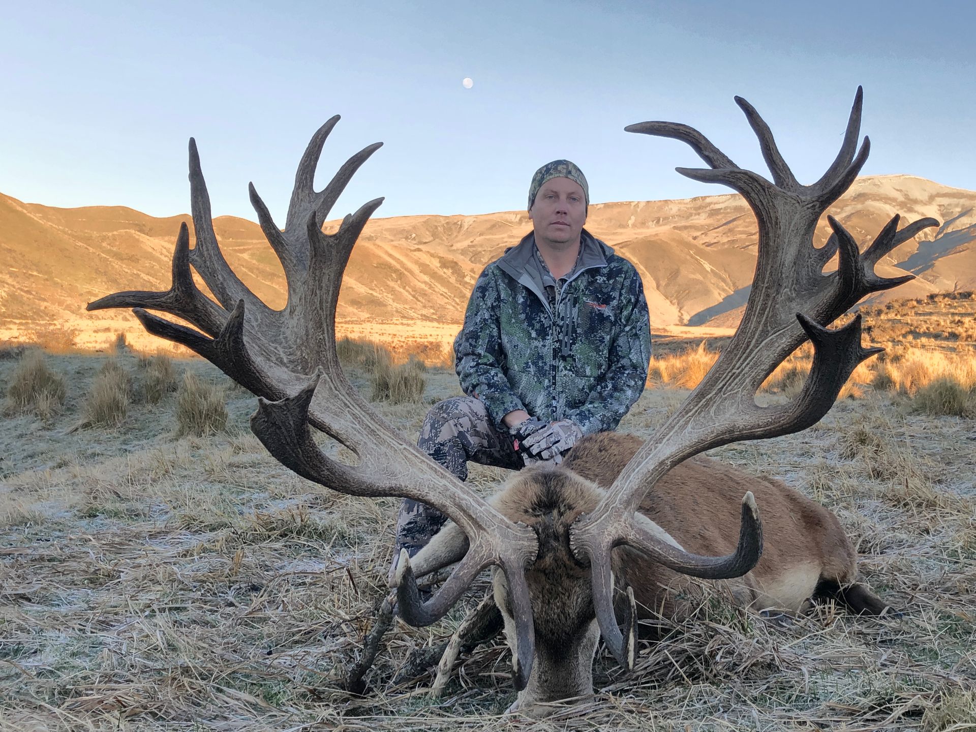 A man in camouflage kneels behind a large red deer stag with massive antlers in a field. Mountains are in the background.