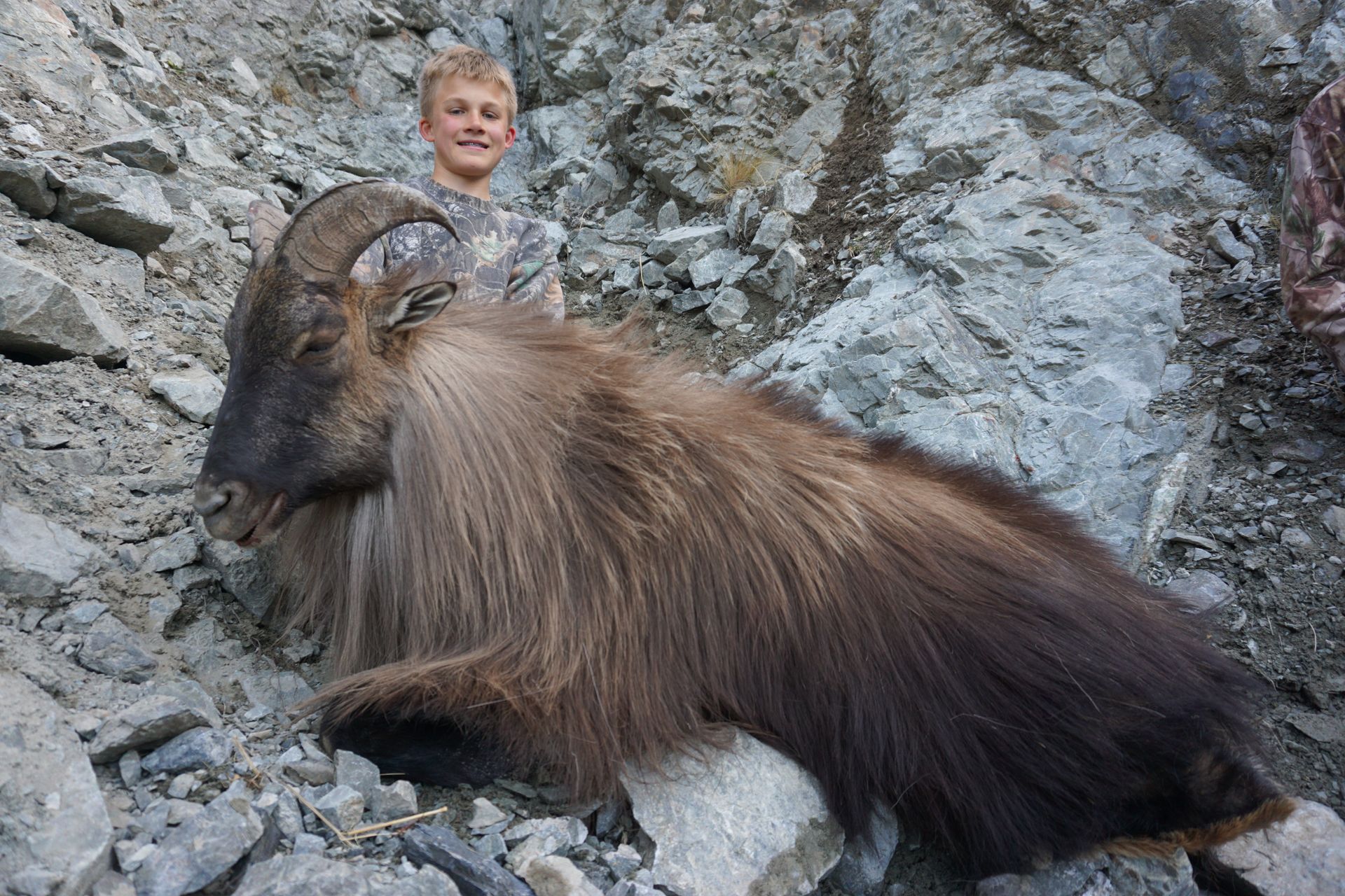 Boy stands next to a large, shaggy-haired Himalayan tahr on a rocky mountainside. The boy smiles; the tahr lies still.