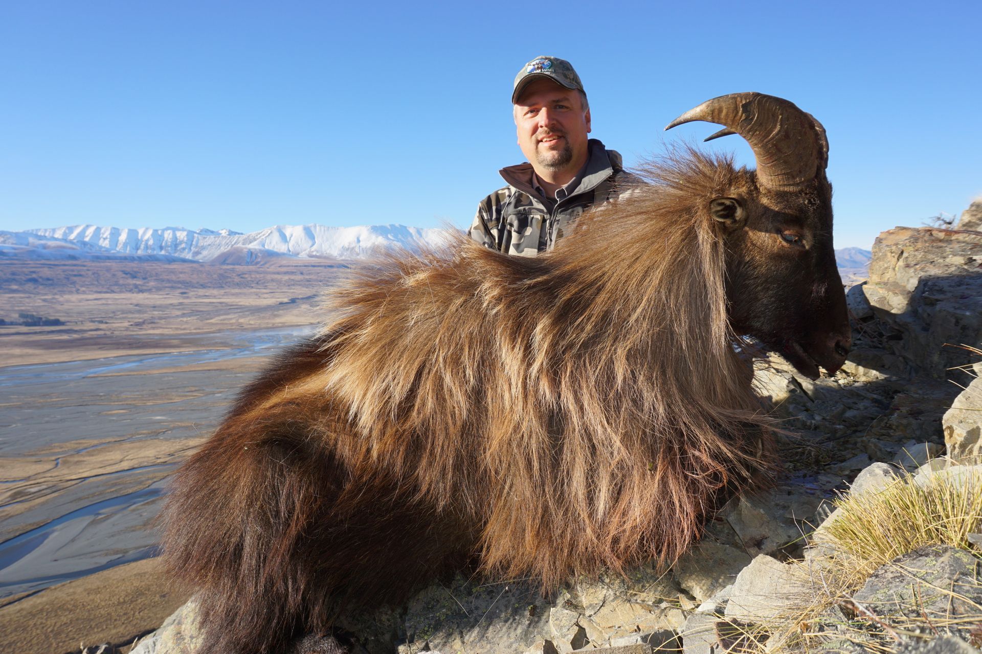 Man in camo holding a dead Himalayan tahr on a rocky outcrop with mountains and valley in the background.