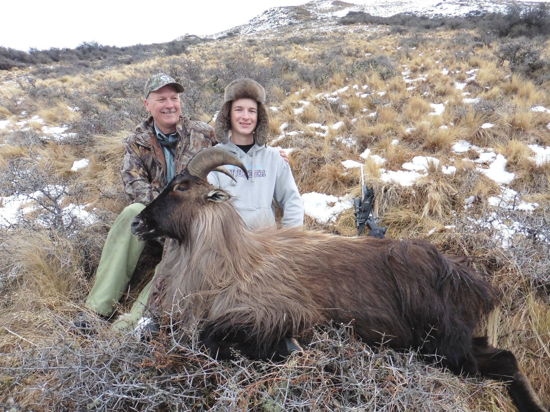 Two people pose with a dead brown Himalayan tahr on a hillside. The man smiles and the woman is wearing a fur hat.