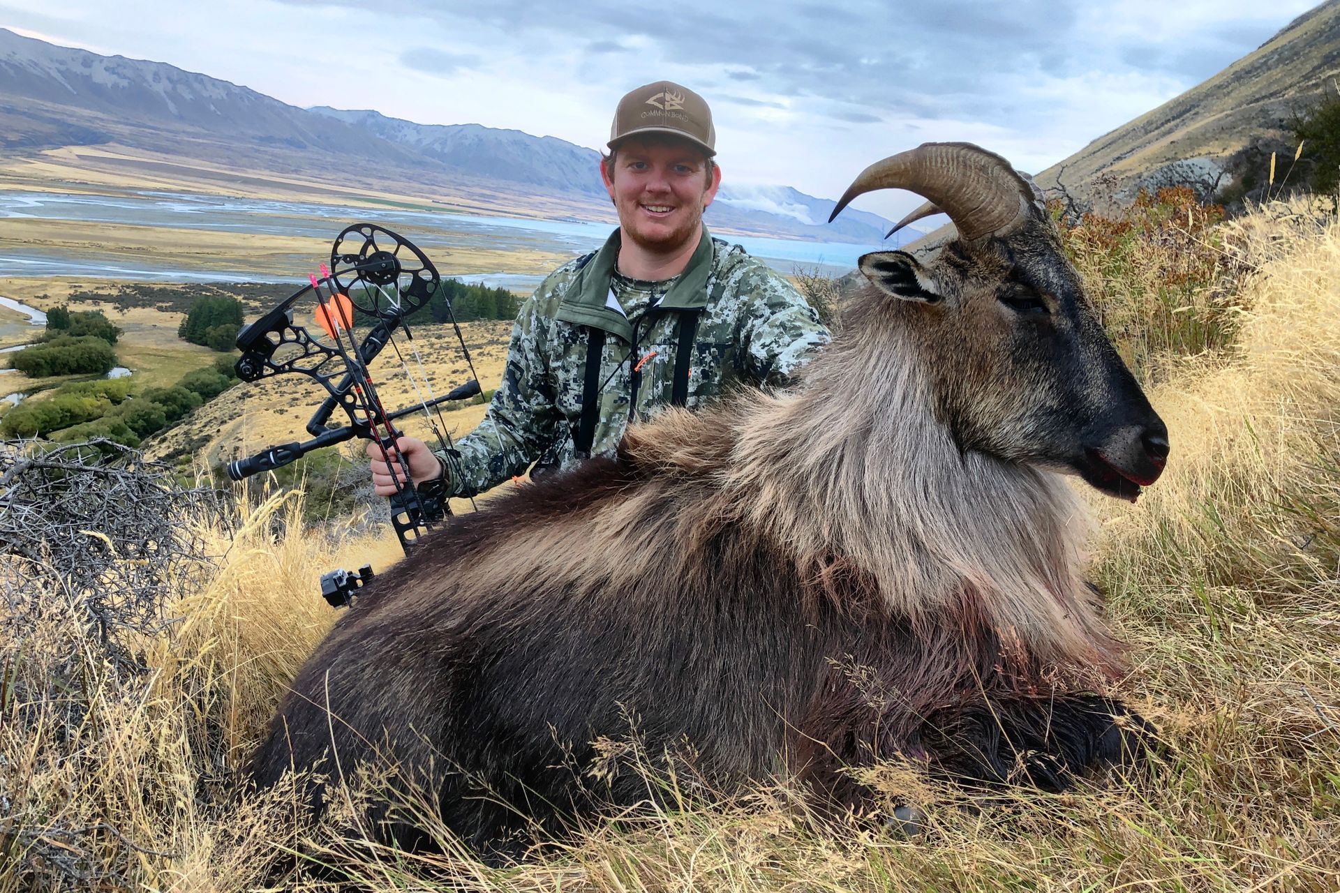 Man in camouflage holding a bow, posing with a large dead Himalayan tahr in a grassy mountain landscape.