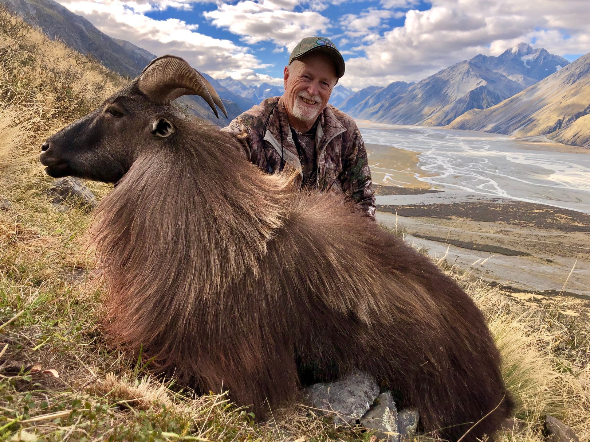 Man in camo with a large, brown Himalayan Tahr in mountainous terrain. The man smiles, posing next to the animal.
