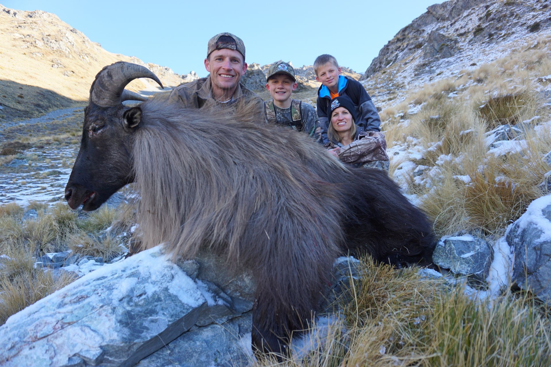 A family poses with a Himalayan tahr on a snowy mountainside. They are smiling, dressed in hunting gear, with the tahr resting on rocks.