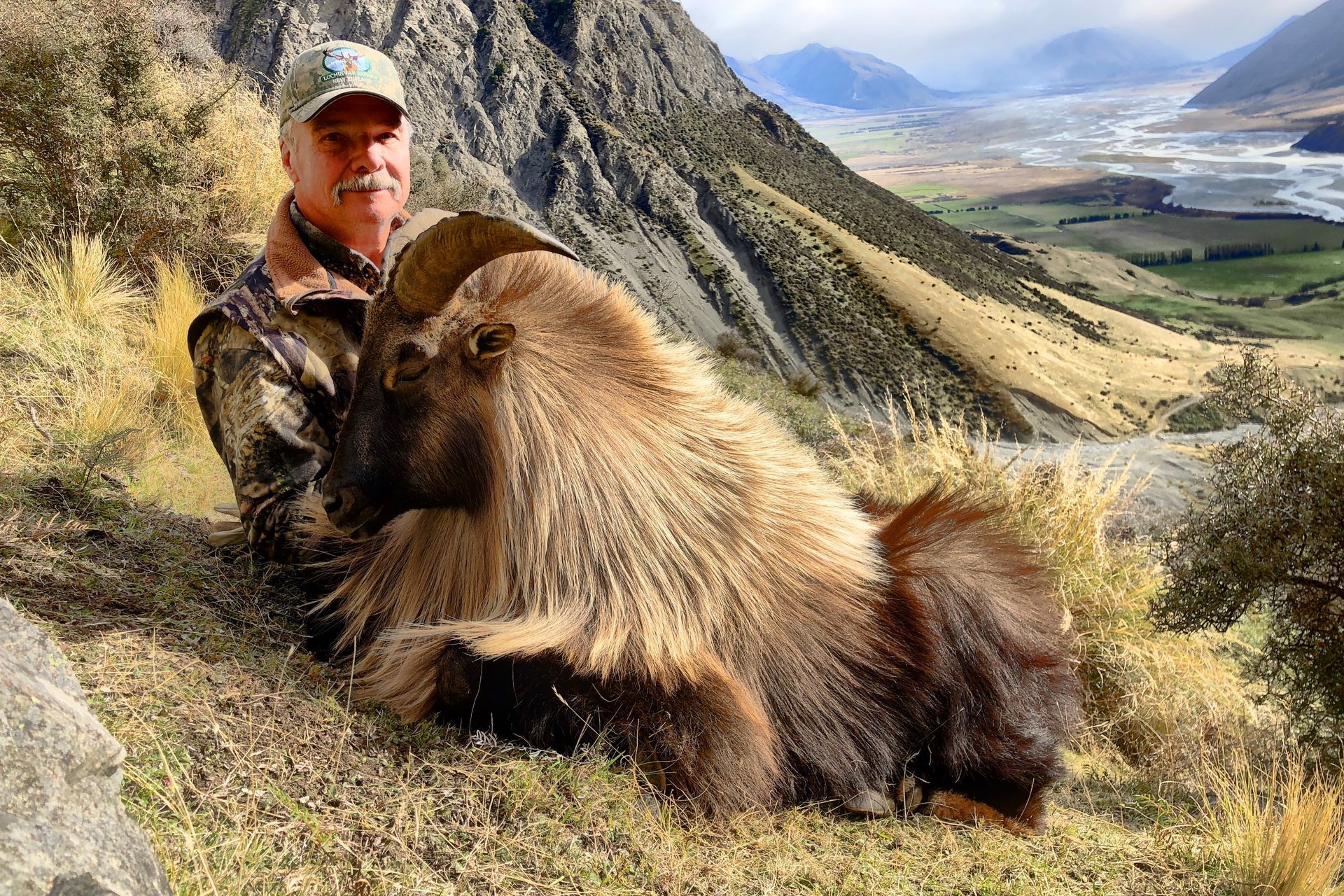 Man in camo holds a large, shaggy, brown animal in a mountainous, grassy landscape.