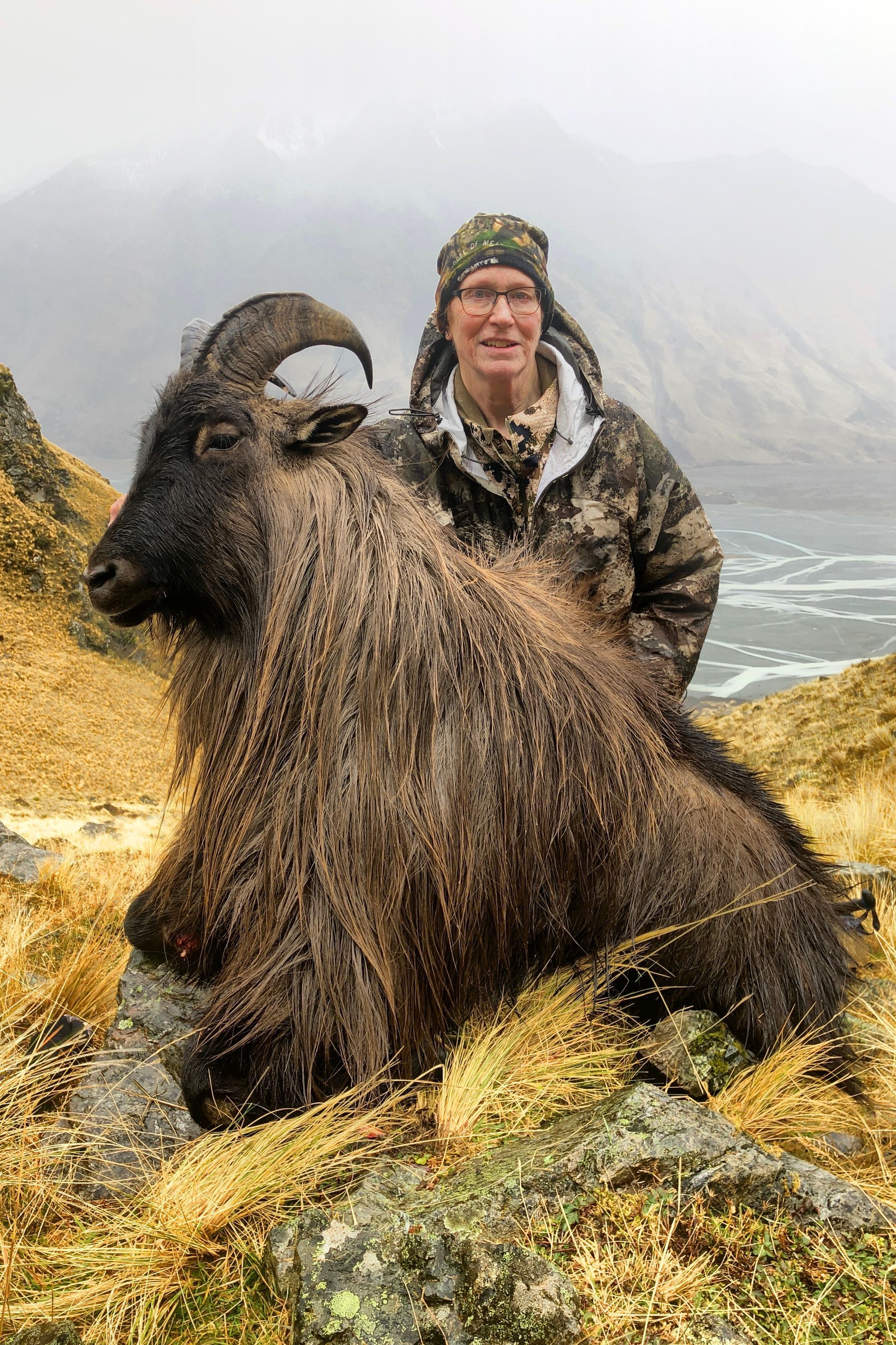 Man in camouflage poses with a large, shaggy brown Himalayan tahr on a rocky, grassy hillside.  Overcast, possibly near a body of water.