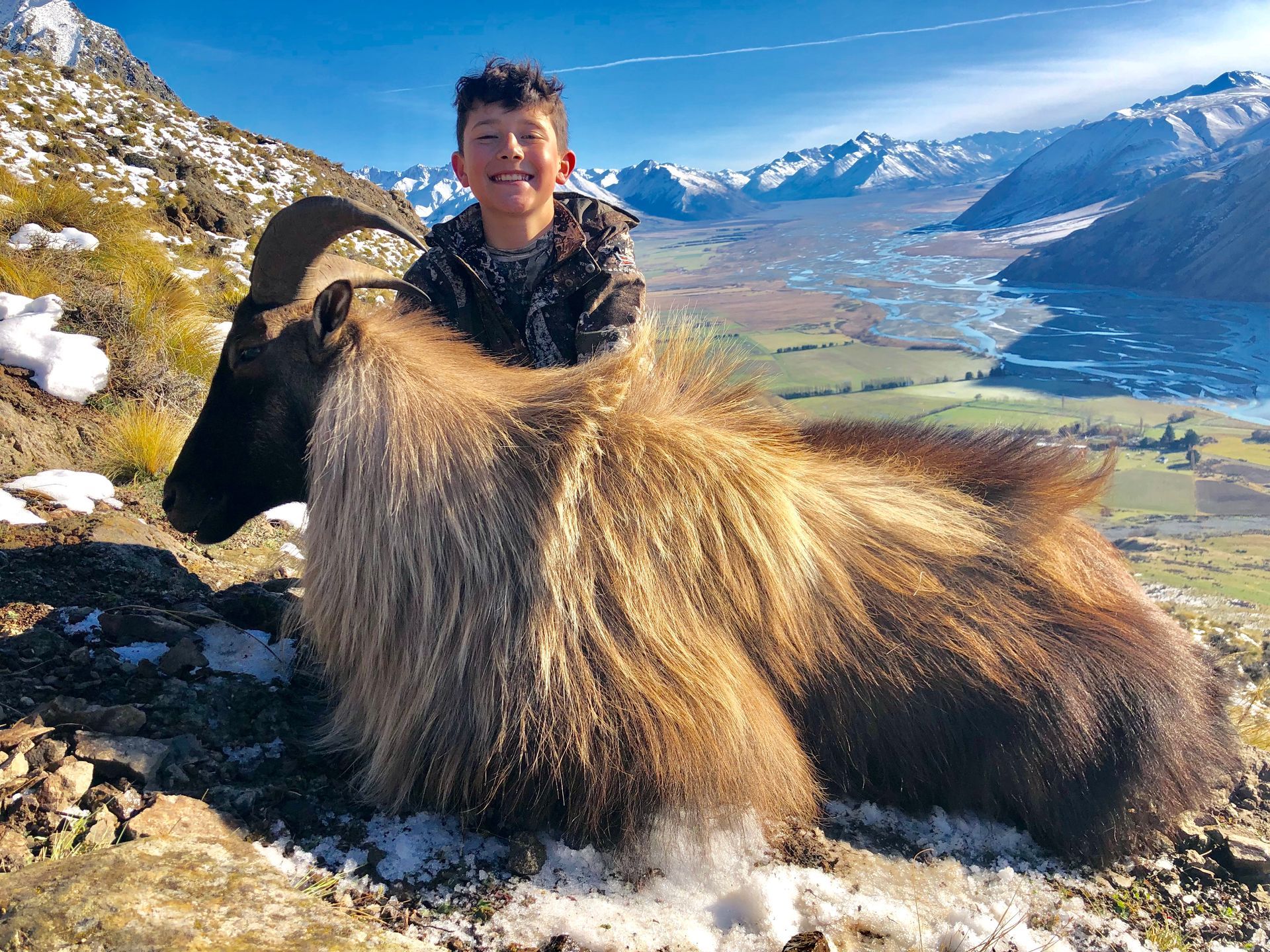 Boy smiles, sitting atop a recently harvested chamois in a snowy mountain landscape. Green valley and mountains in the distance.