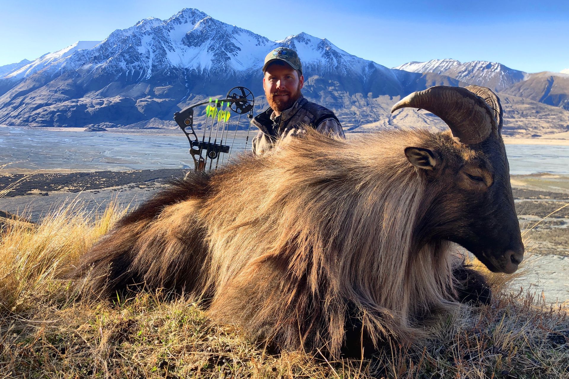 Man with a bow proudly poses with a harvested Himalayan Tahr in a mountainous landscape. Brown fur contrasts with a snowy backdrop.