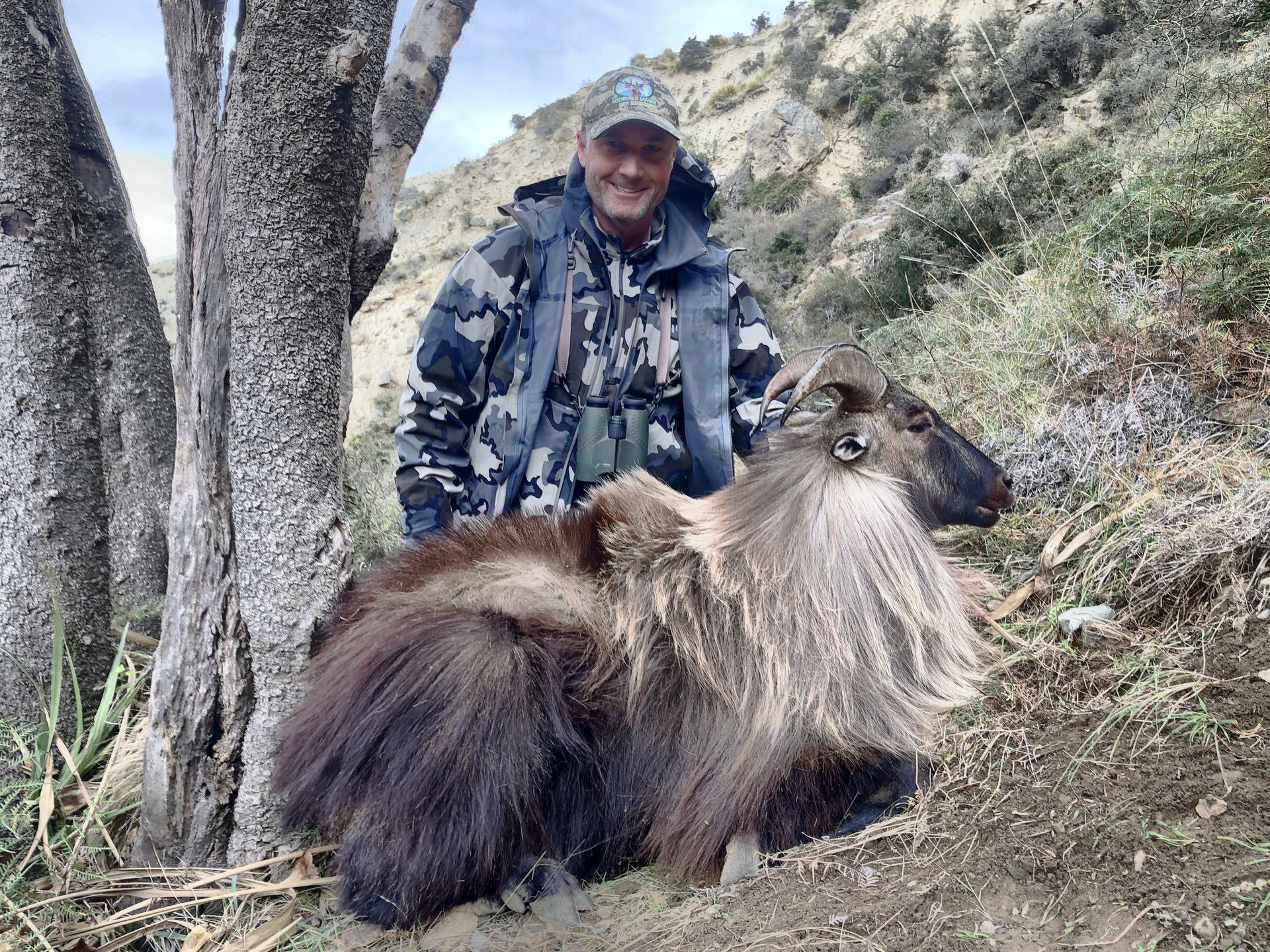 Man in camouflage with a Himalayan Tahr he hunted in a mountainous environment. He is smiling near a tree.