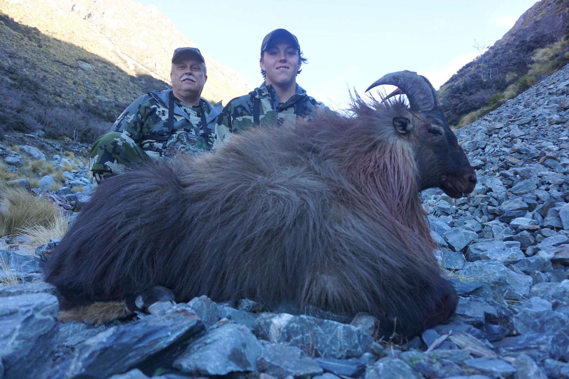 Two hunters pose with a large, dead Himalayan tahr in a rocky mountain landscape. The tahr has long, dark brown fur.