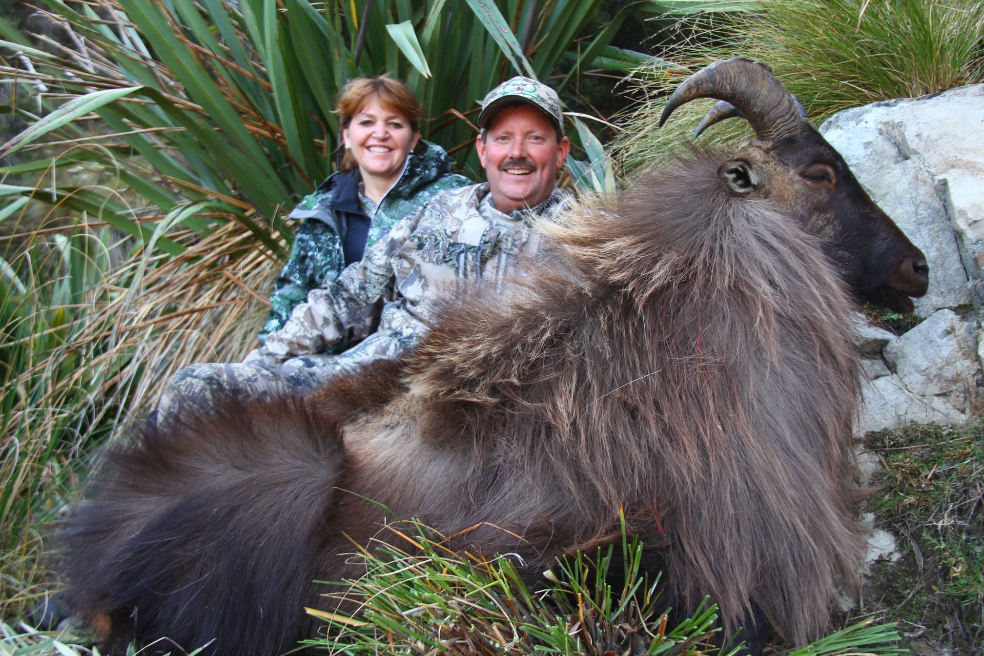A man and woman in camouflage pose with a large, shaggy animal they appear to have hunted outdoors. They smile at the camera.