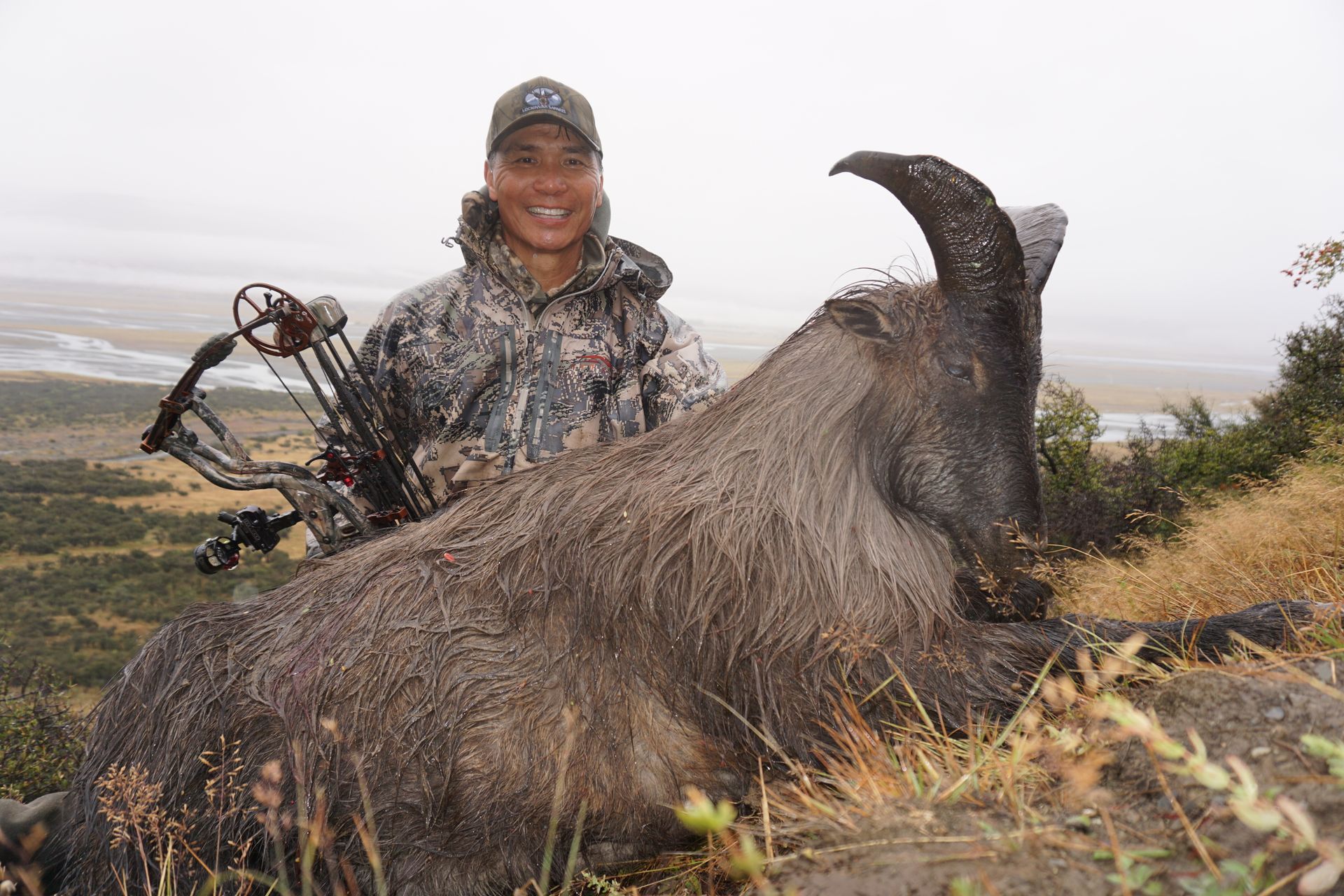 Hunter in camo with a bow, smiling next to a large, dark-colored muskox on a hillside, with a river visible in the background.