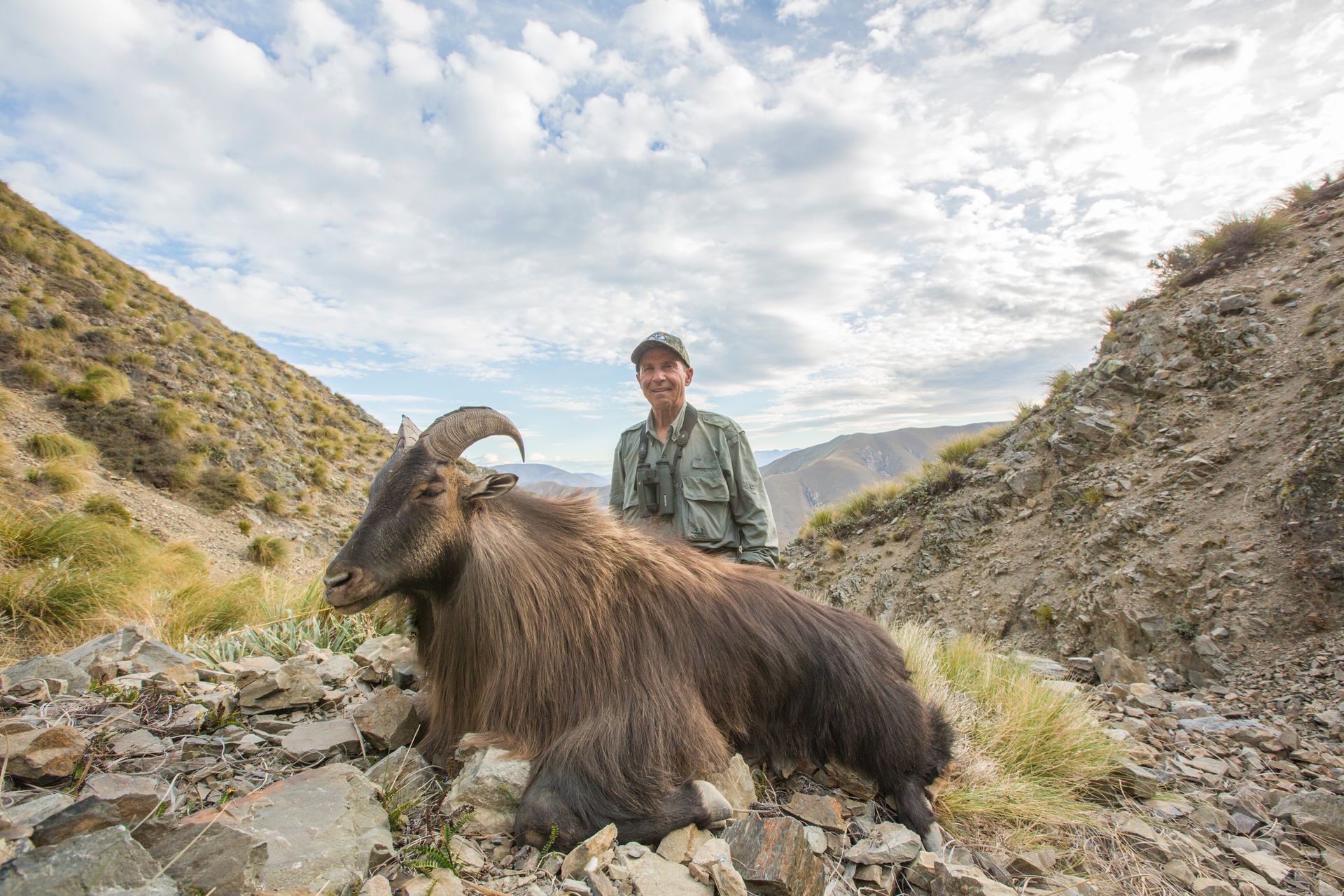 Man posing with a large, brown Himalayan tahr he hunted in a rocky mountain landscape.
