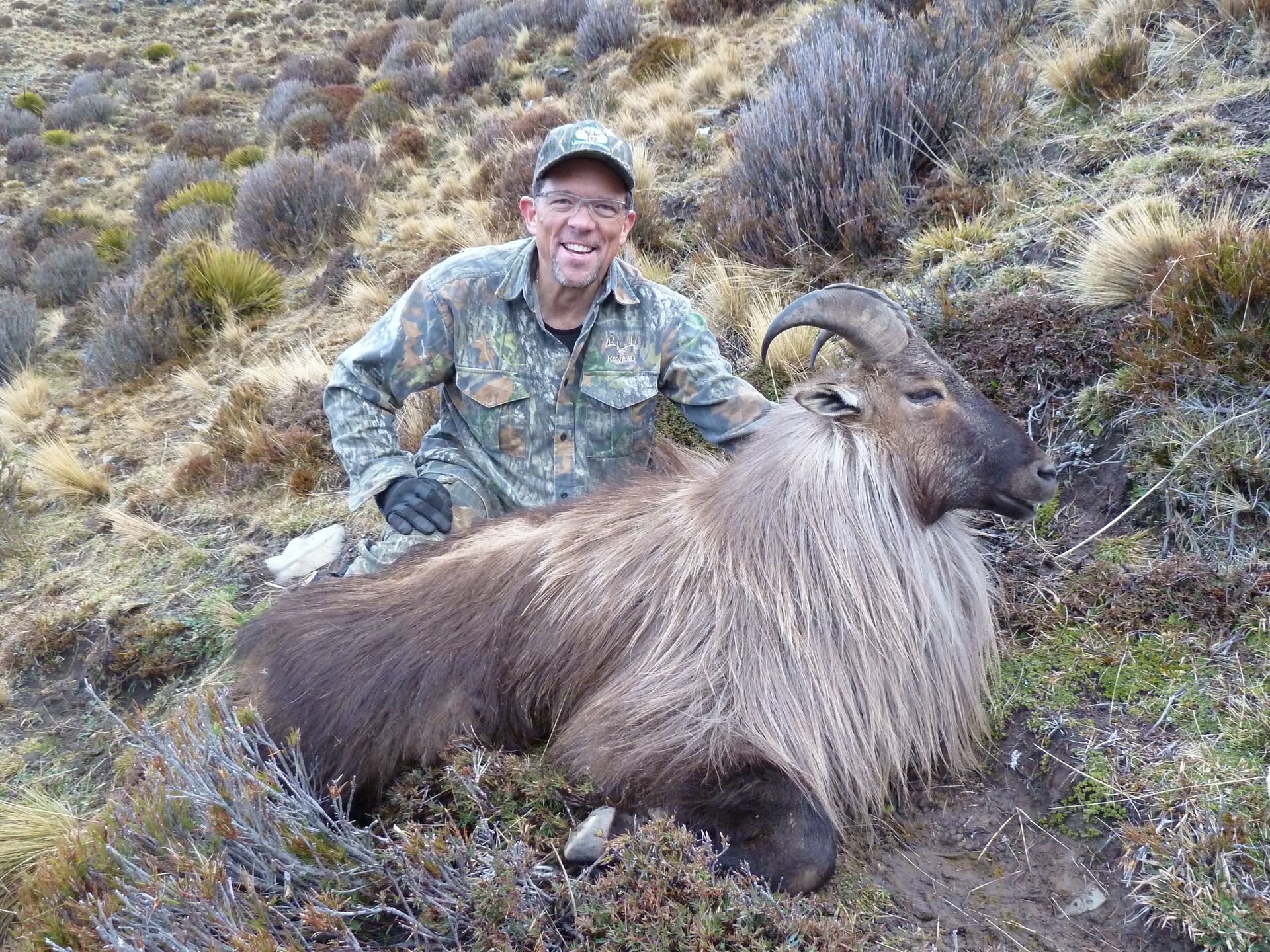 Man in camouflage next to a Himalayan Tahr. They're on a hillside. The man is smiling, and the Tahr has a long, shaggy mane.