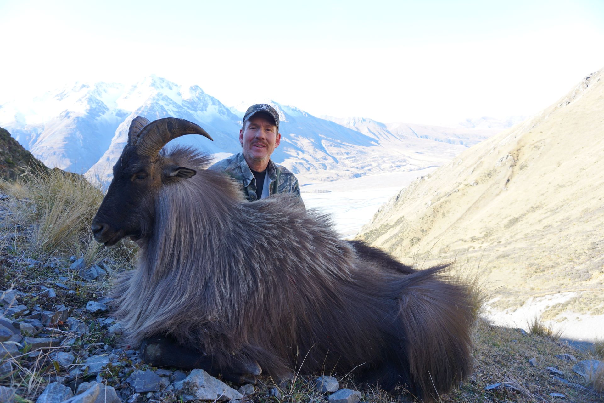 Hunter poses with a large, shaggy Himalayan Tahr in a mountainous landscape. Snow-capped peaks are visible in the background.
