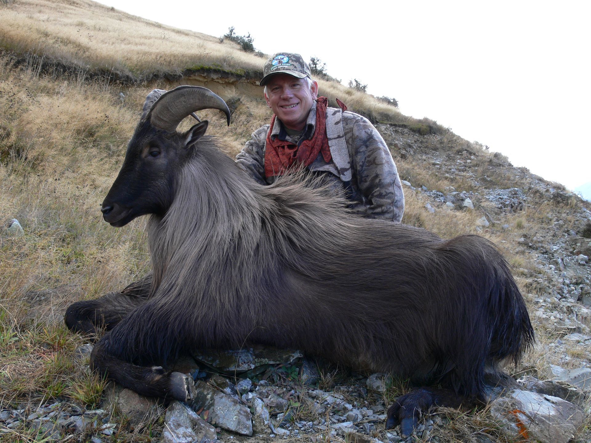 Man in camouflage gear posing with a Himalayan tahr he appears to have hunted, on a hillside with dry grass.