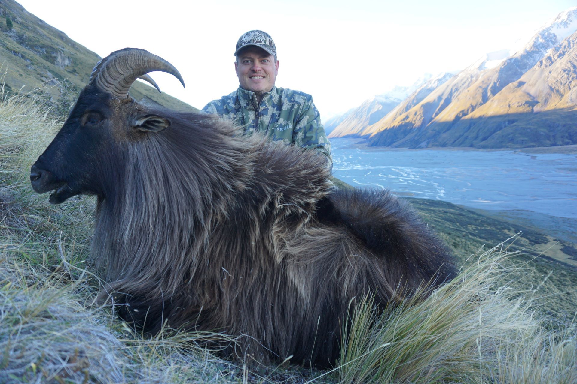 Man poses with a large, dark-haired tahr in a mountainous, grassy landscape; a river and mountains are in the background.