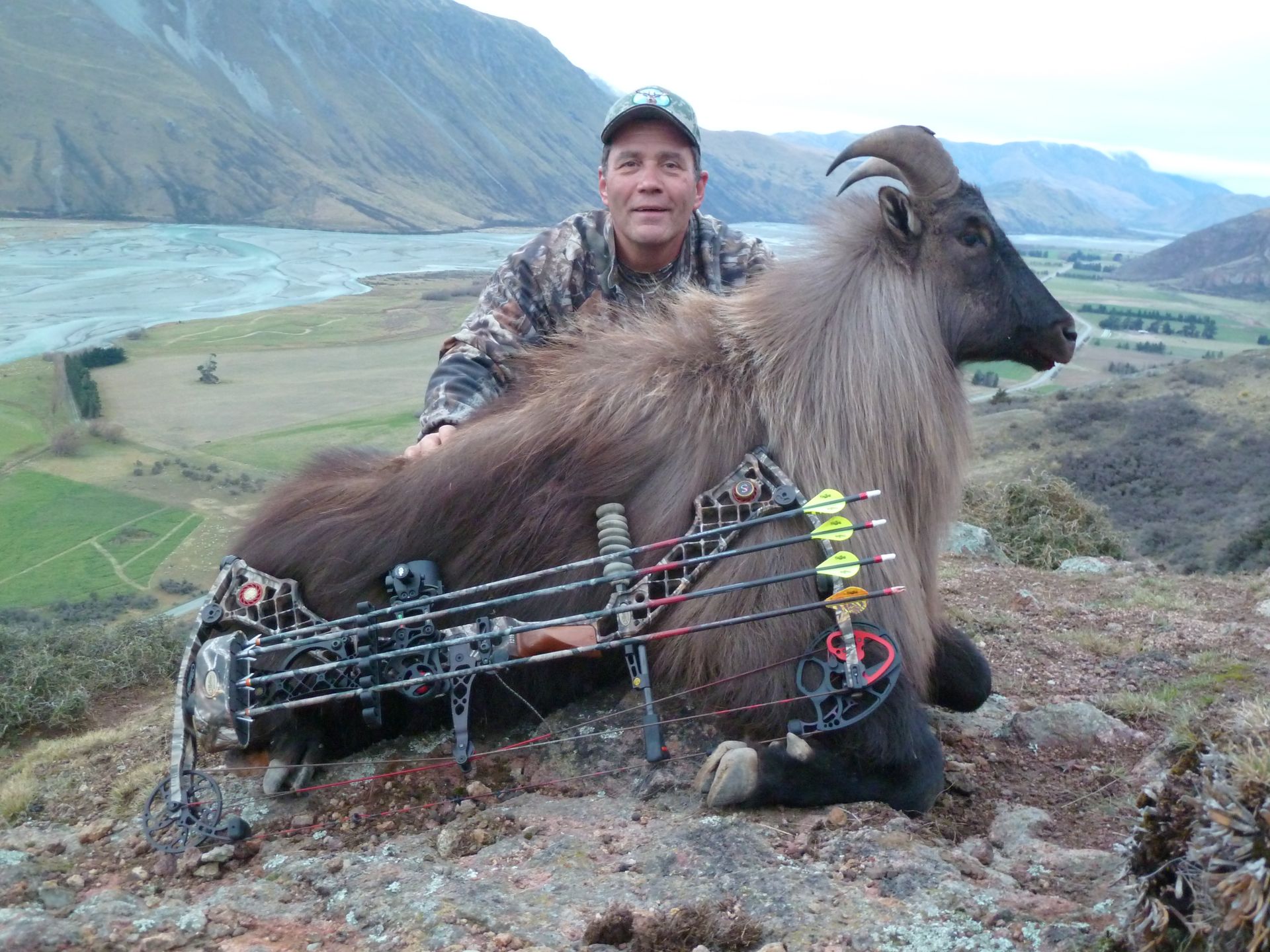 Hunter in camouflage poses with a Himalayan tahr and a compound bow on a hillside overlooking a river.