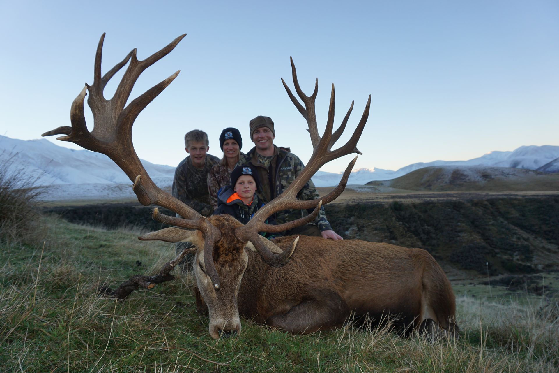 Four people pose with a large, dead deer with impressive antlers in a grassy field, mountains in the background.