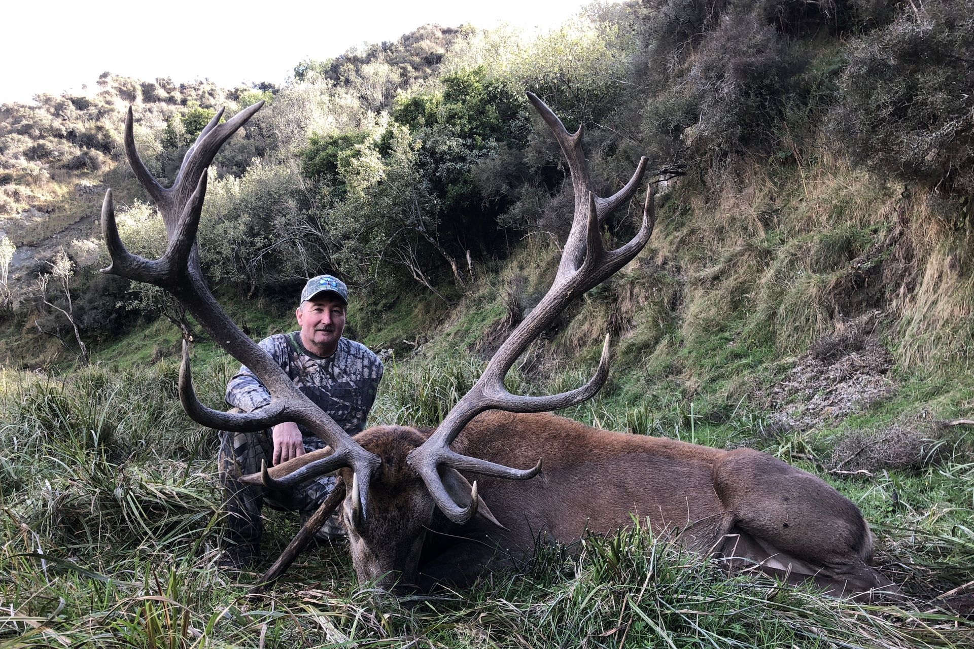 Man kneeling beside a large dead red deer with impressive antlers, in a grassy, hilly environment.