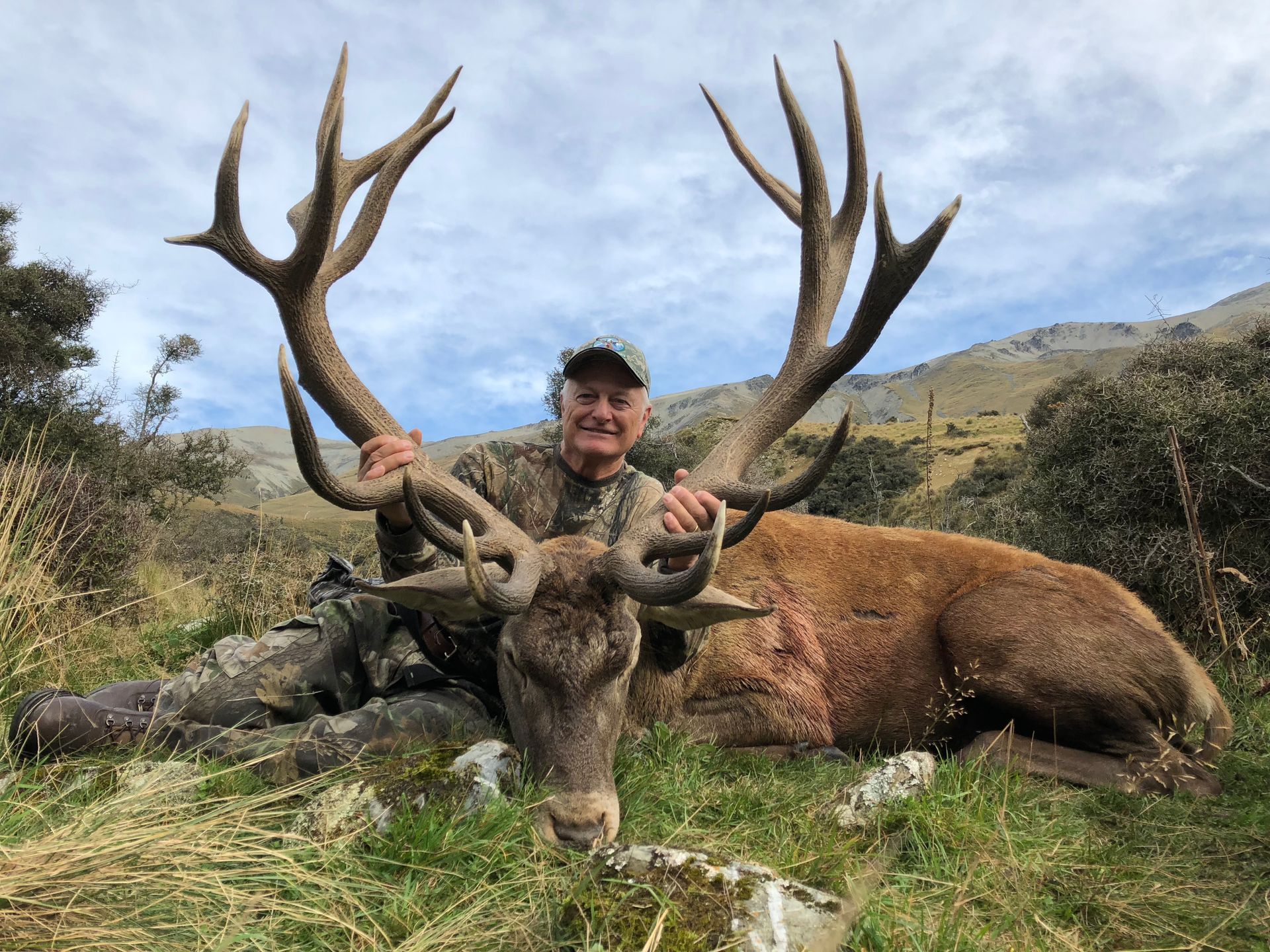 Man in camouflage smiles, holding a large deer's antlers; deer lies on grass with mountains in the background.