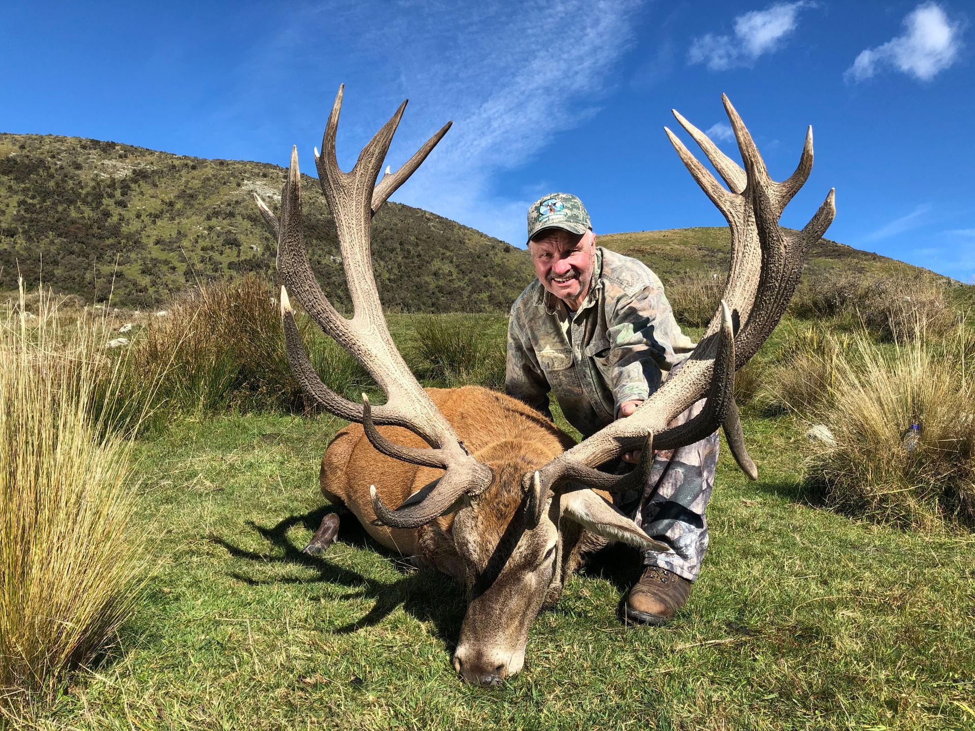Hunter smiles, posed beside a large dead deer with massive antlers, in a grassy field with hills in the background under a blue sky.