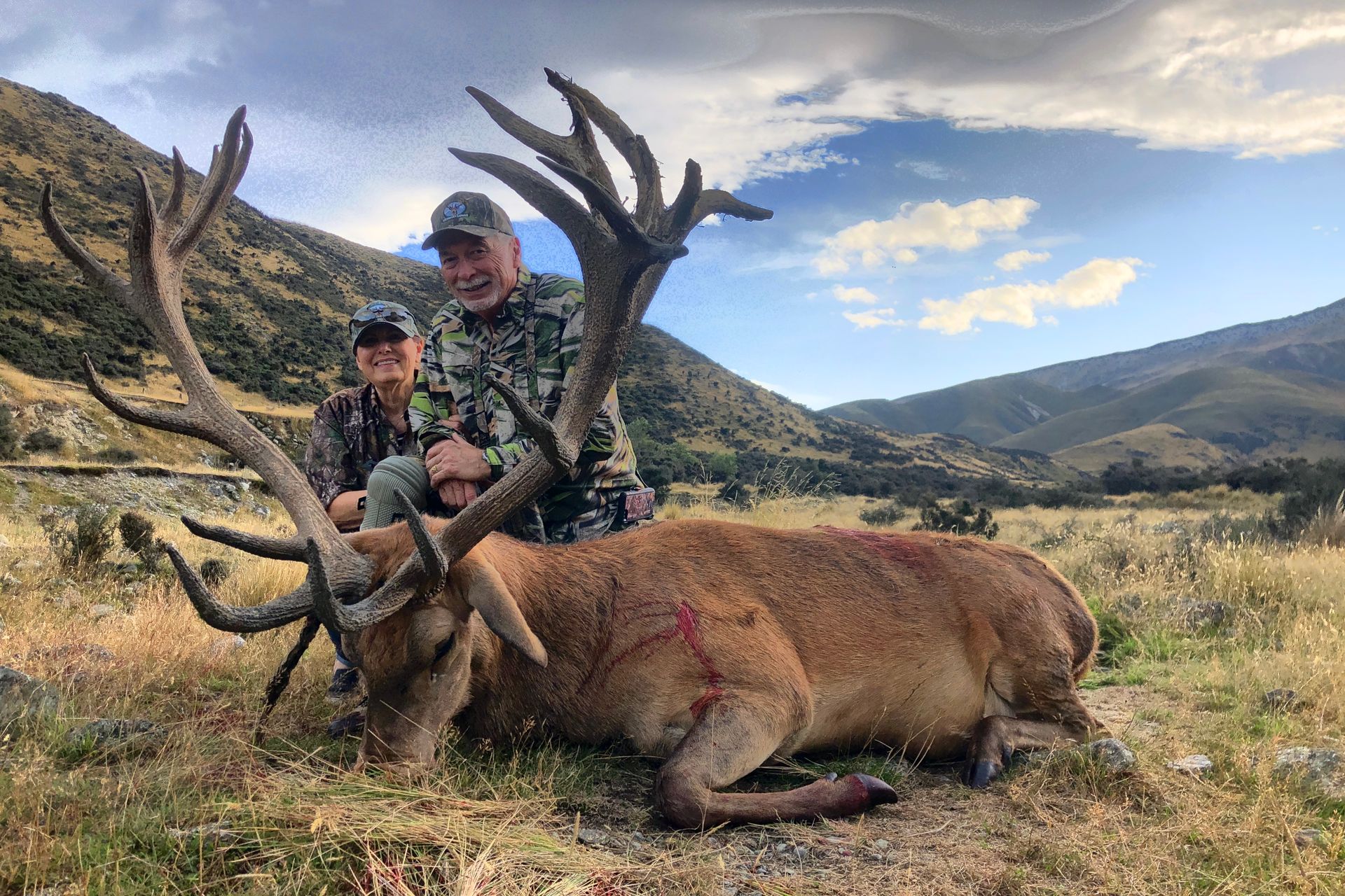 Two hunters pose with a large, dead red deer in a mountainous landscape under a blue sky with clouds.