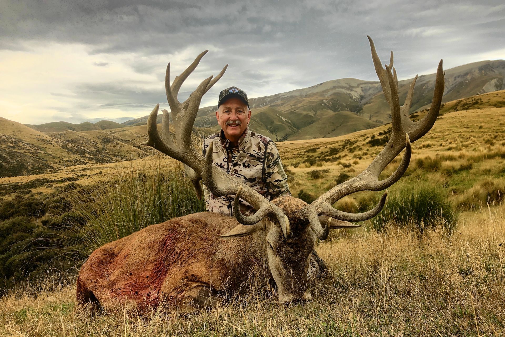 Man in camouflage smiling, posing with a large dead stag with massive antlers, set in a grassy field and hills.