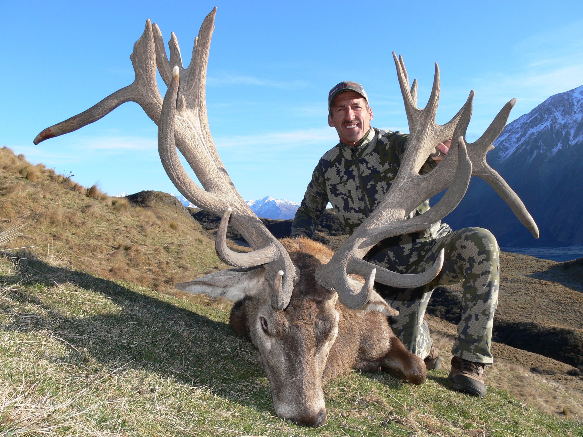 Man kneels beside a large red deer stag with impressive antlers, outdoors in a mountainous setting.