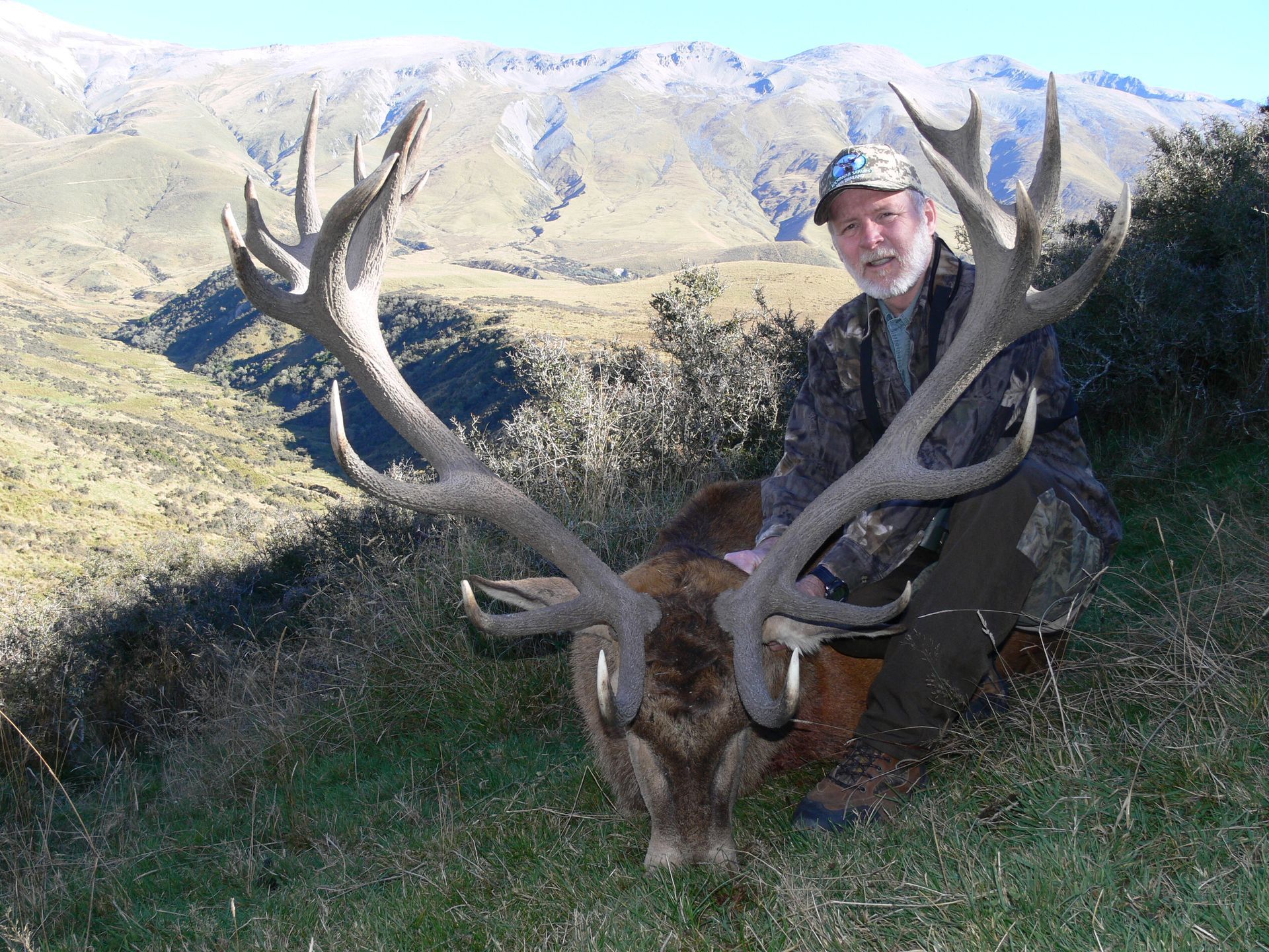 Man in camouflage kneeling beside a large, dead deer with impressive antlers. Hills and mountains are in the background.