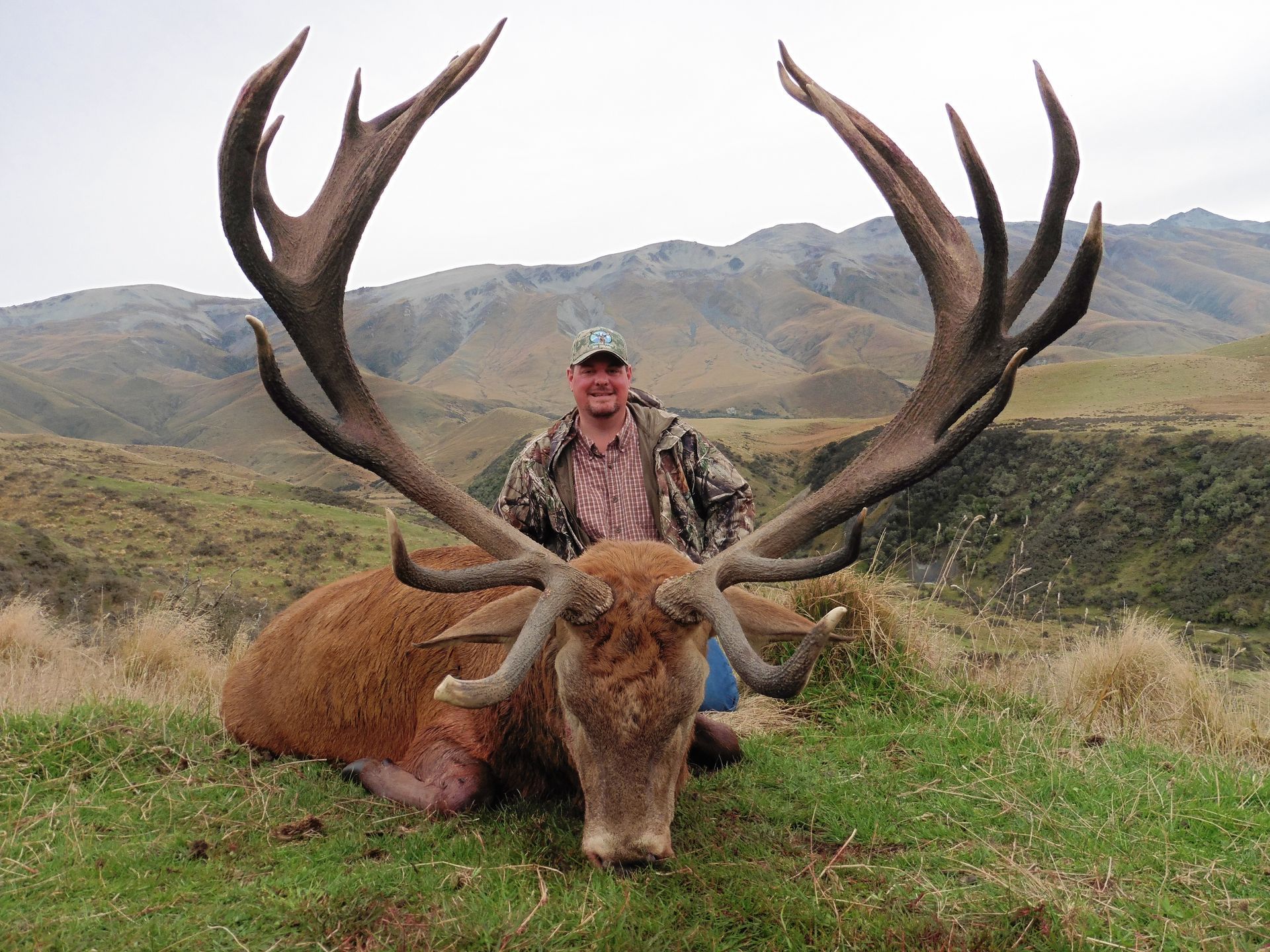 A man in camouflage kneels beside a large red deer with massive antlers in a grassy, mountainous landscape.