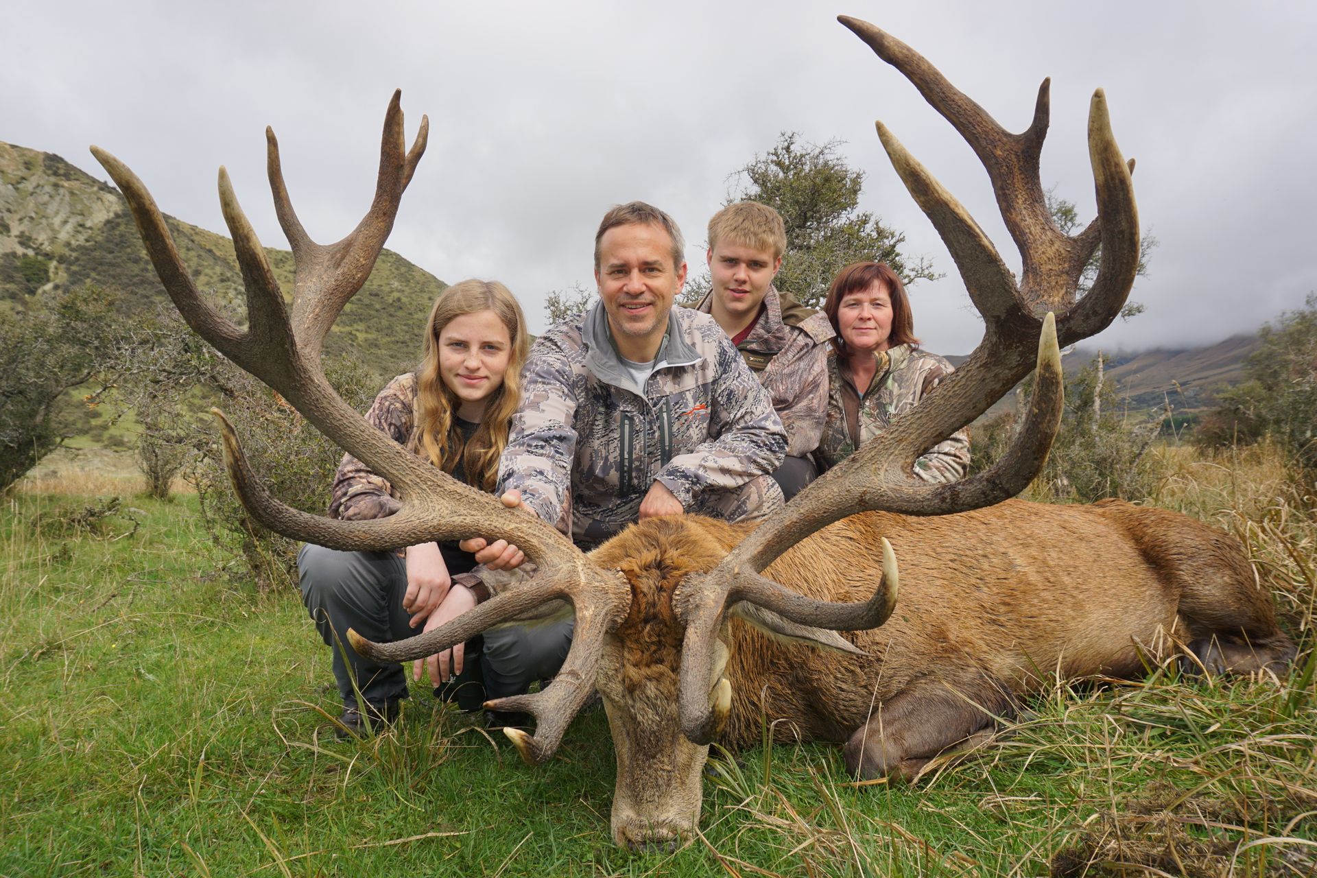 A family poses with a large dead red deer they've hunted. They are in a grassy, outdoor setting.
