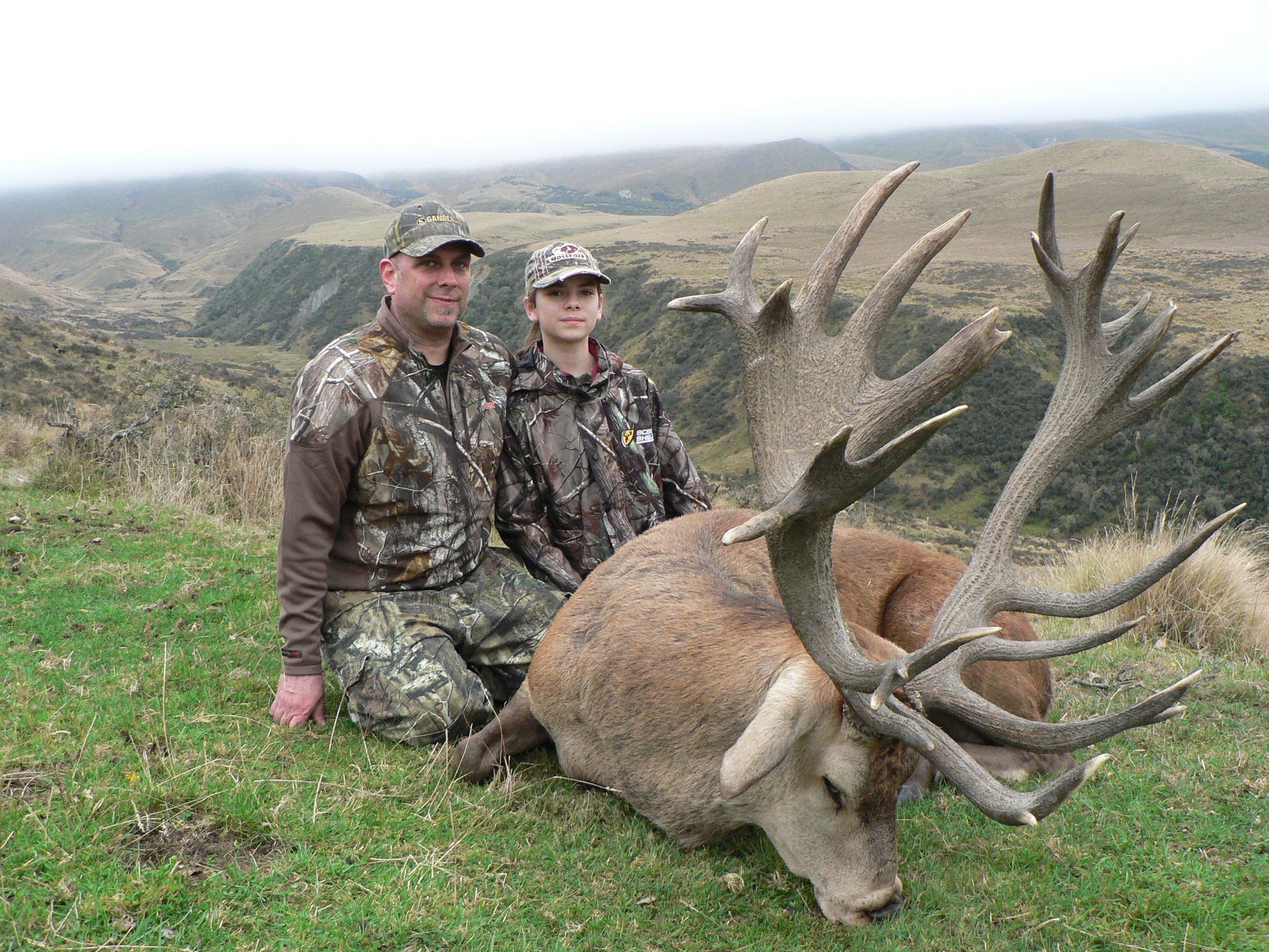 Man and young person in camouflage pose with a large red deer stag in a hilly, grassy setting.