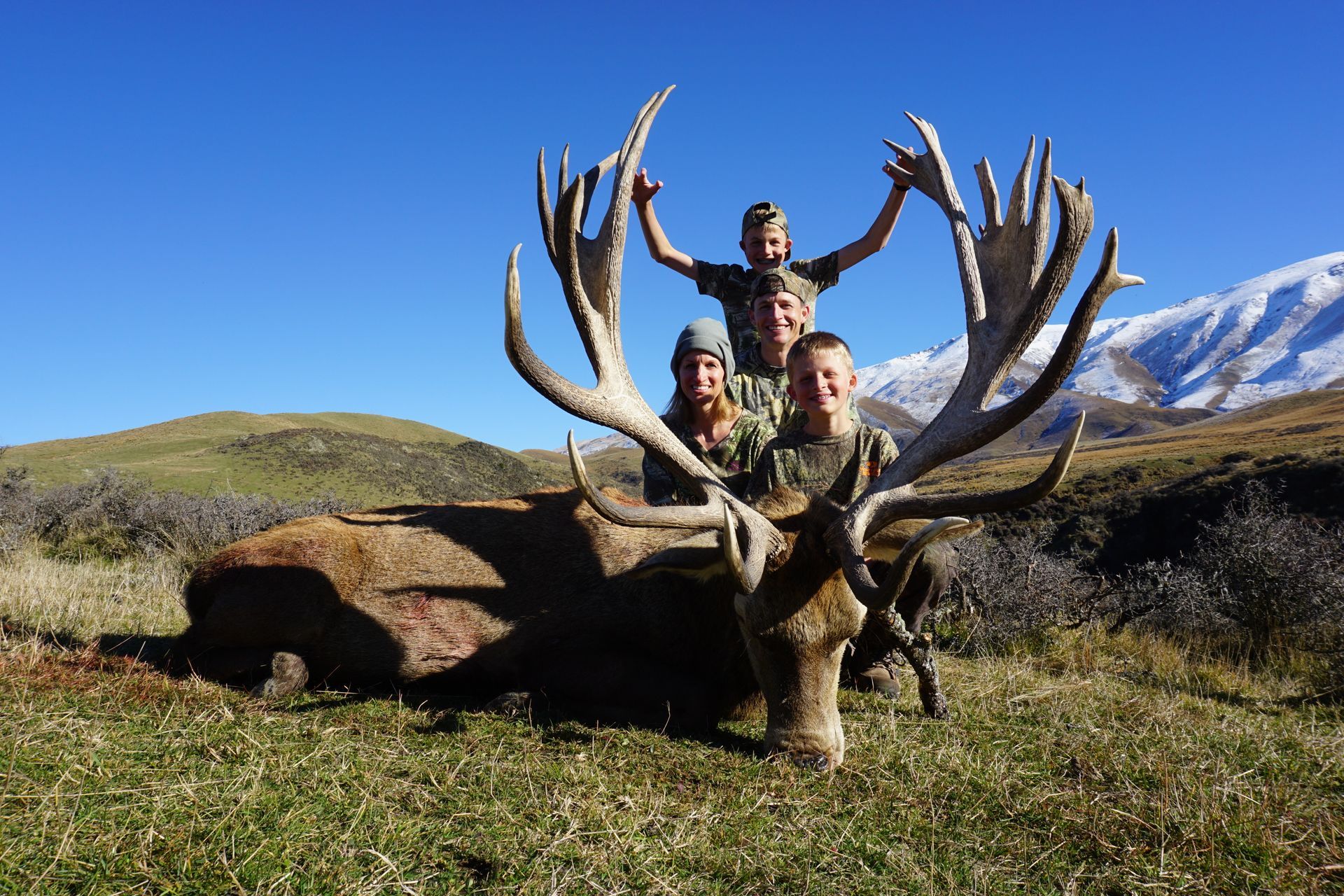 Three people pose with a large dead deer in a grassy field with mountains in the background.