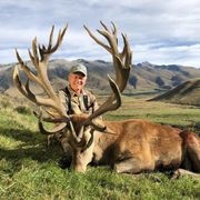 A man in camouflage smiling next to a large dead red deer with impressive antlers in a grassy, mountainous landscape.