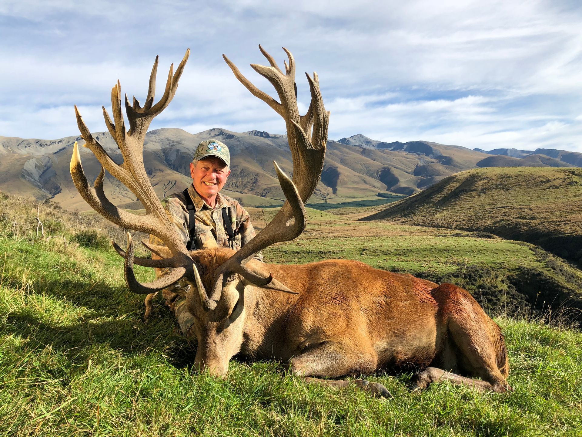 Man in camouflage poses with a large dead red deer buck in a grassy field with mountains in the background.