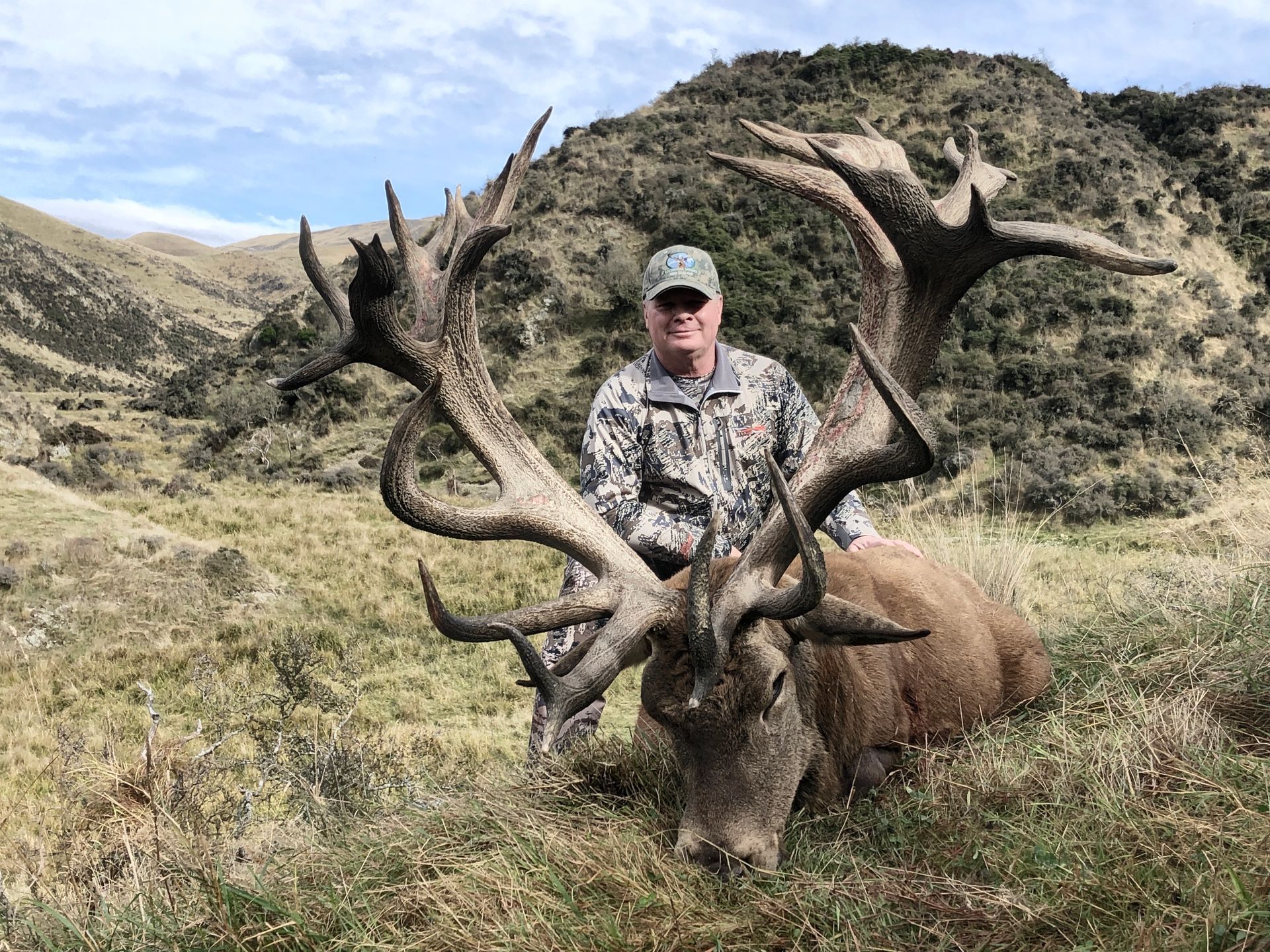 Man in camouflage with a large dead stag, its antlers spanning wide in a grassy mountain setting.