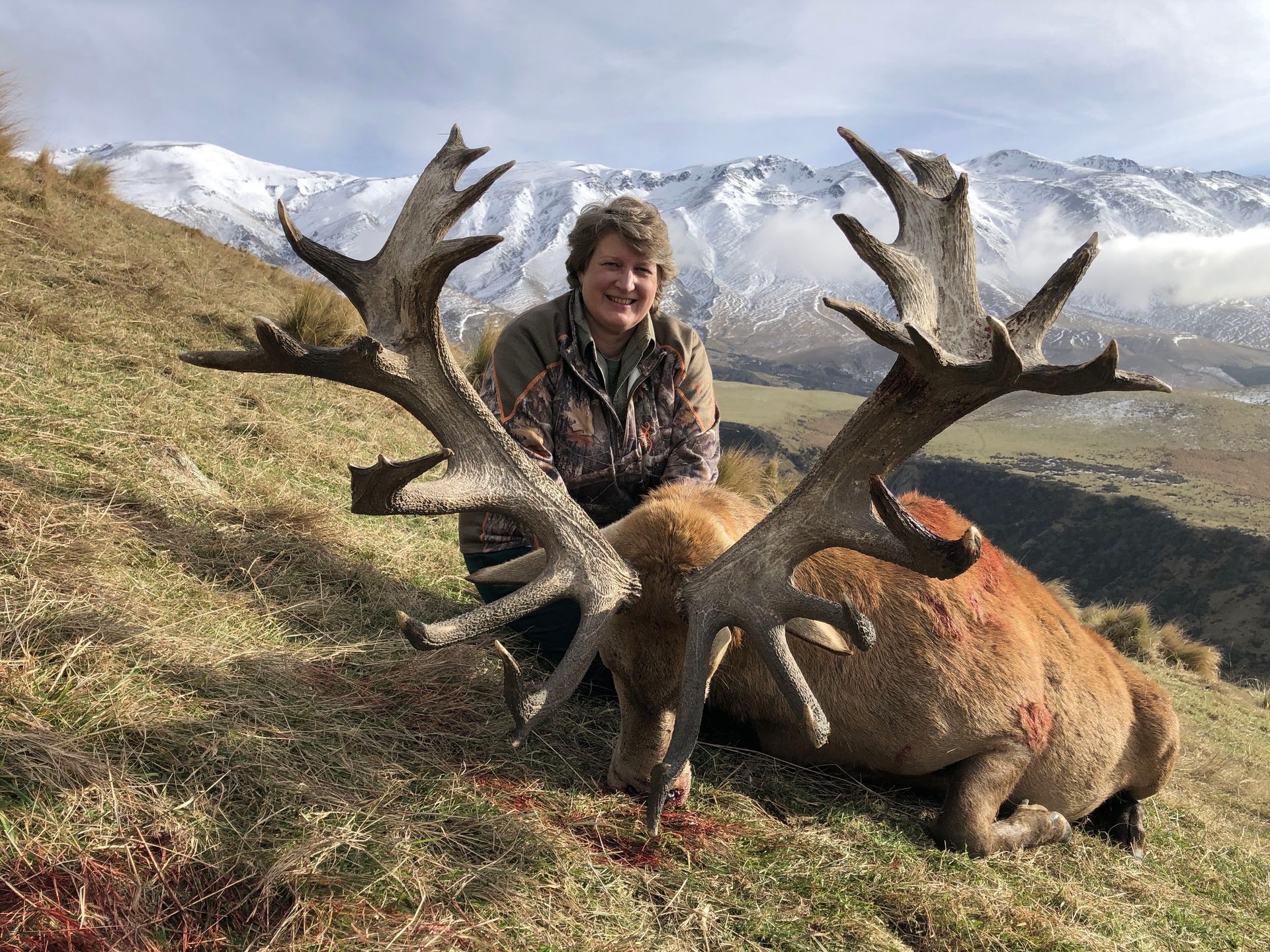 A woman kneels next to a large, dead deer with impressive antlers on a grassy hillside with snow-capped mountains in the background.